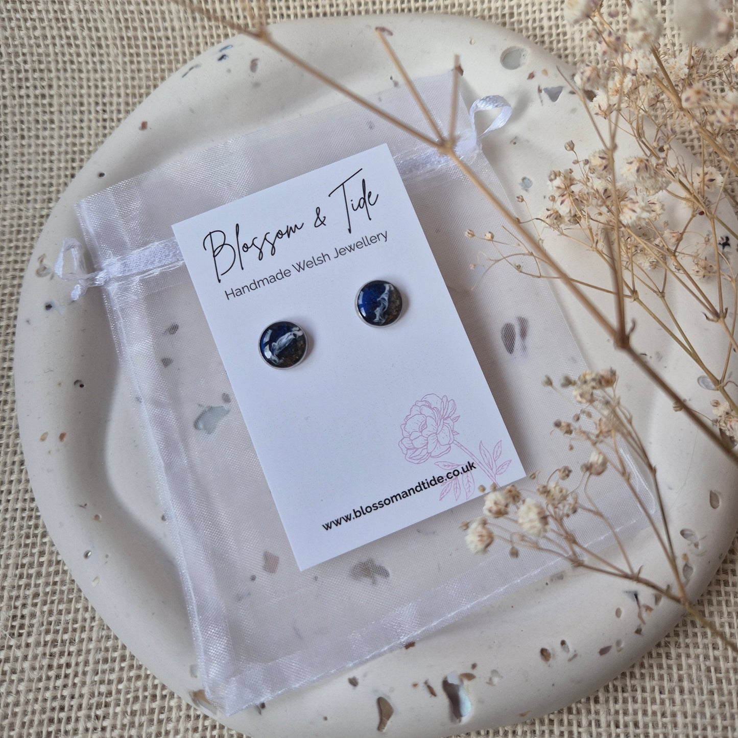 Pair of beach sand earrings on a card with a decorative stone and dried flowers on a textured fabric background