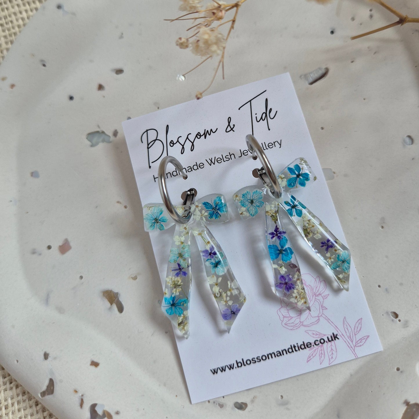 Bow earrings filled with a mix of blue and white pressed flowers on a stainless steel silver hoop. Presented on a Blossom & Tide branded card.
