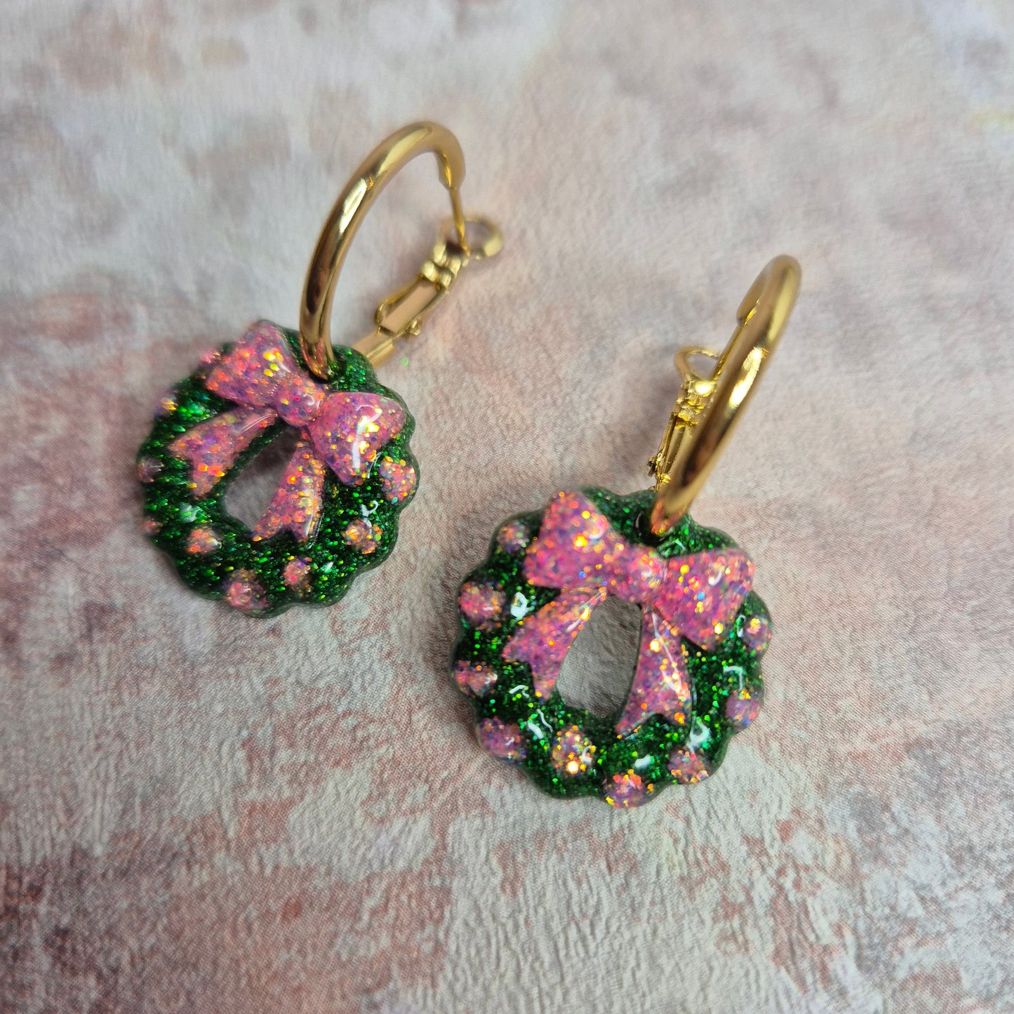 A Close up of a pair of Christmas hoop earrings in green and pink glitter. Wreath and bow design. Against a pink background