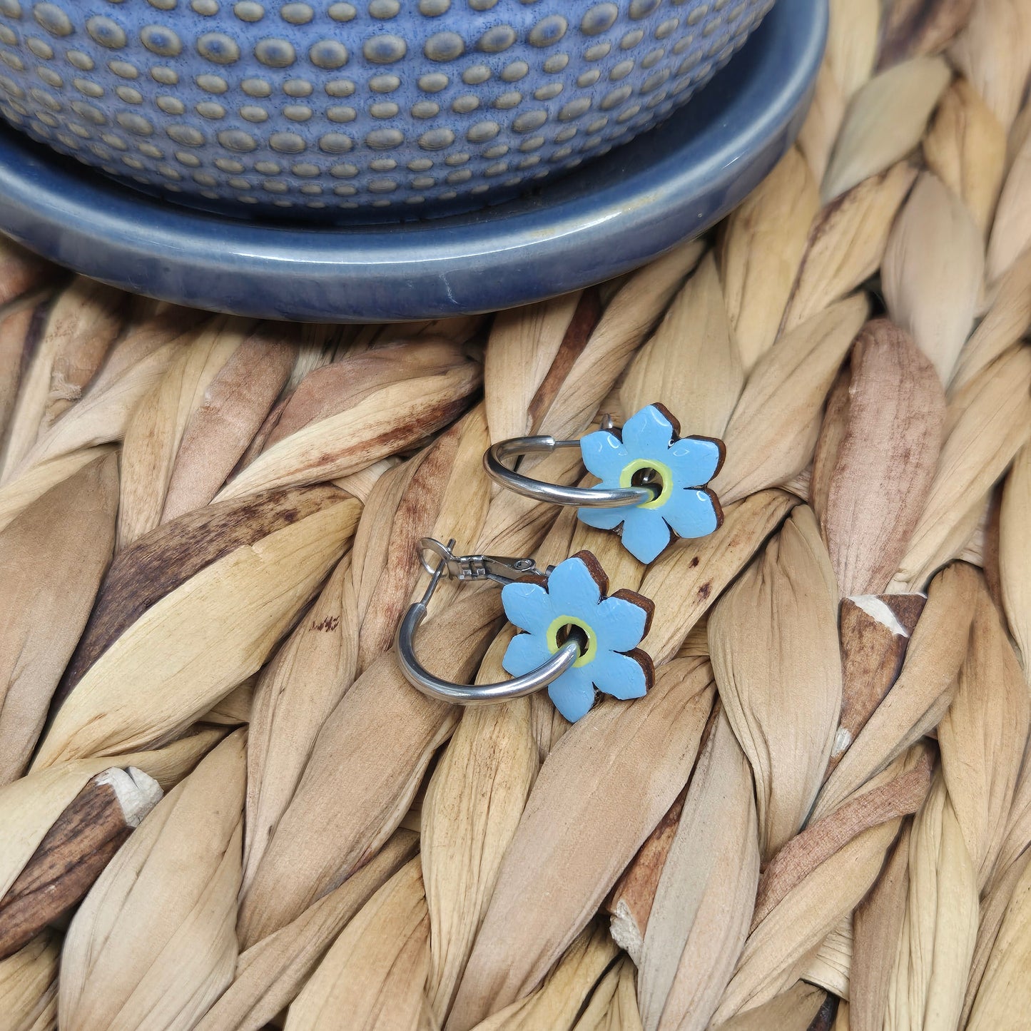 A pair of hand-painted hoop earrings featuring intricate forget-me-not flowers, sealed with a glossy resin topcoat, displayed on stainless steel hoops. Also available in a matte finish.