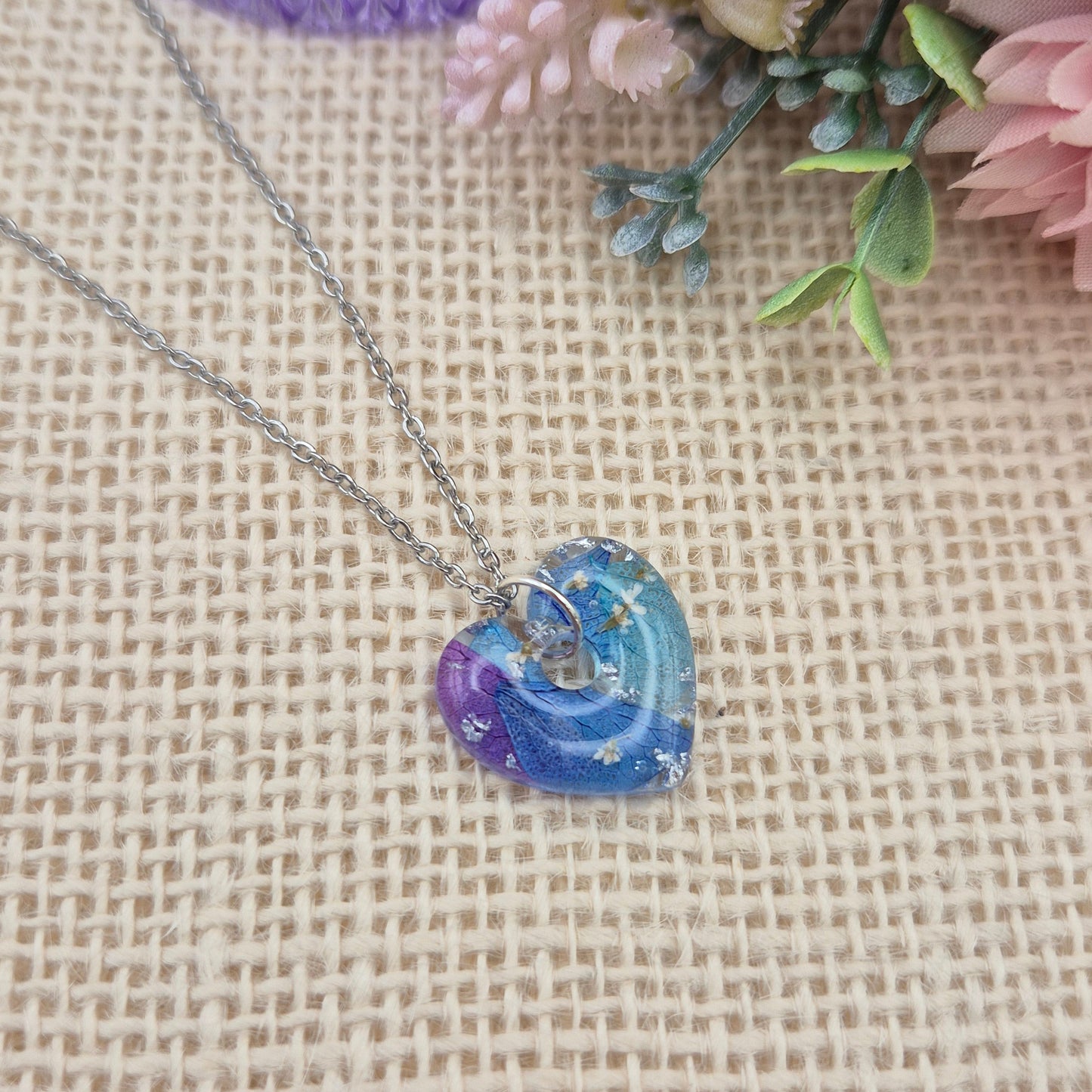 A heart-shaped resin necklace filled with blue and purple hydrangea petals, white flowers, and silver leaf on a silver-coloured stainless steel chain.