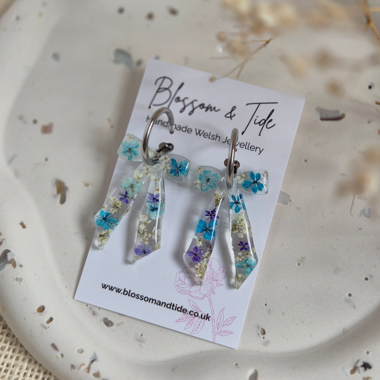 Bow earrings filled with a mix of blue and white pressed flowers on a stainless steel silver hoop. Presented on a Blossom & Tide branded card.