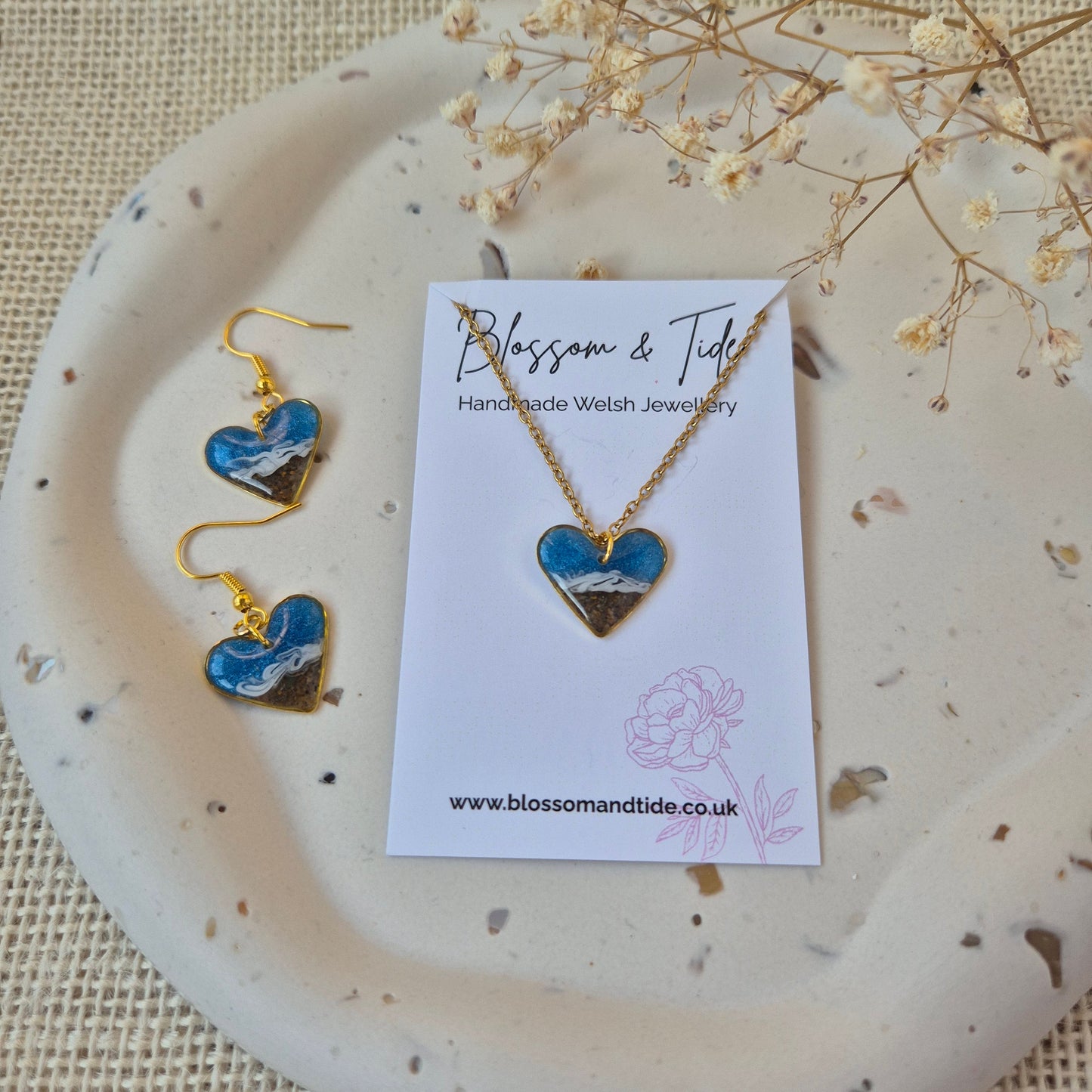 heart shaped sand earrings and matching necklace with gold accents on a branded card, displayed on a textured surface.