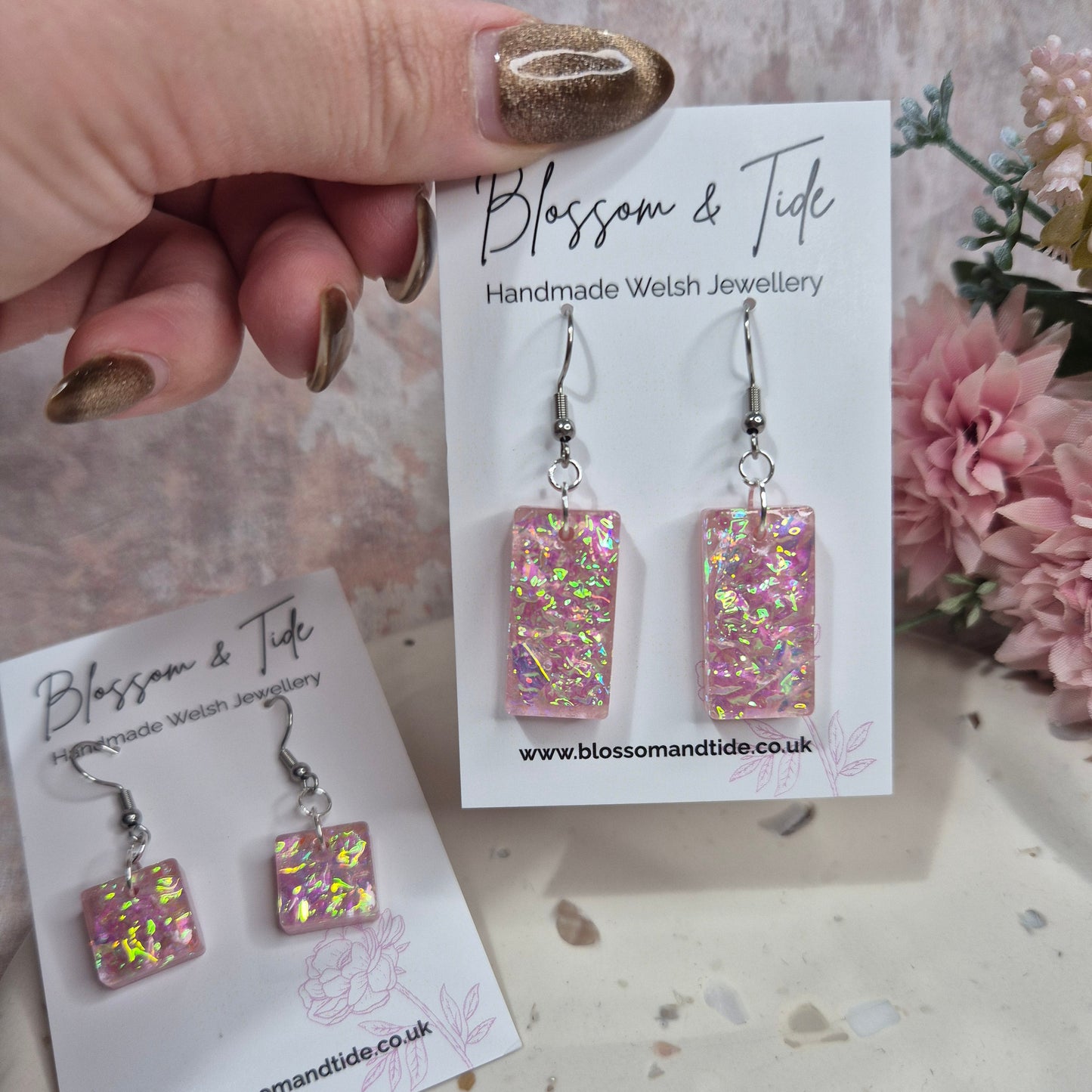 Pink dichroic resin earrings on a 'Blossom & Tide' branded card with flowers in the background.