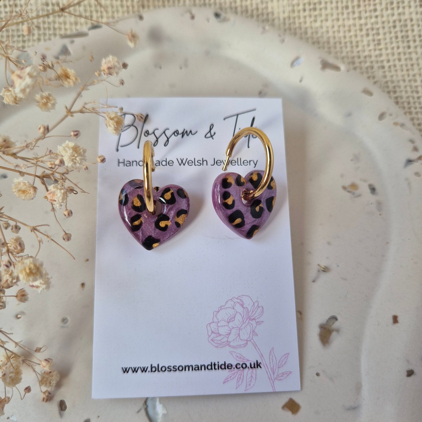 Heart-shaped earrings with gold and purple design on a branded card, placed on a textured surface with dried flowers.