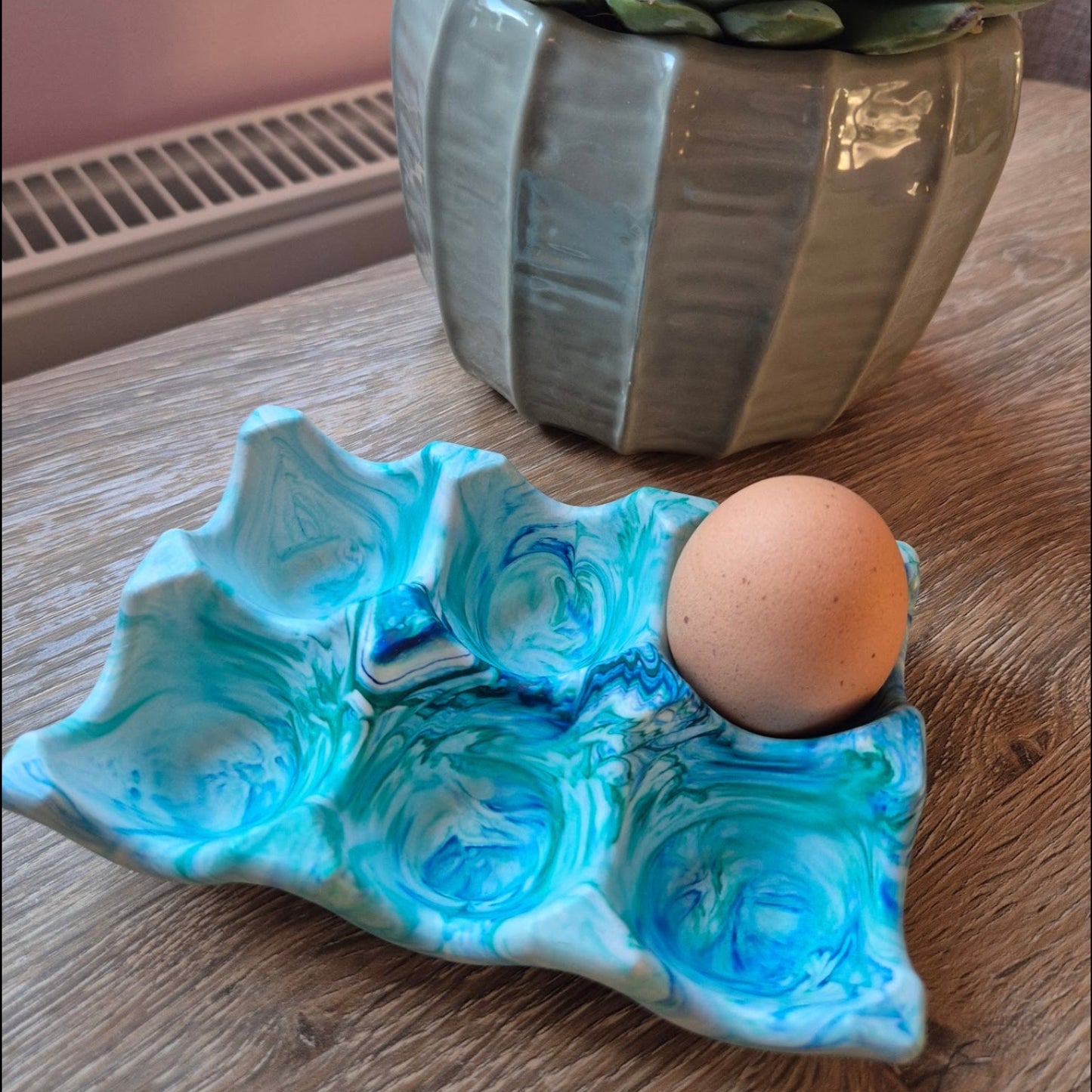 Blue and green Jesmonite egg holder on a wooden surface, with a potted plant in the background.