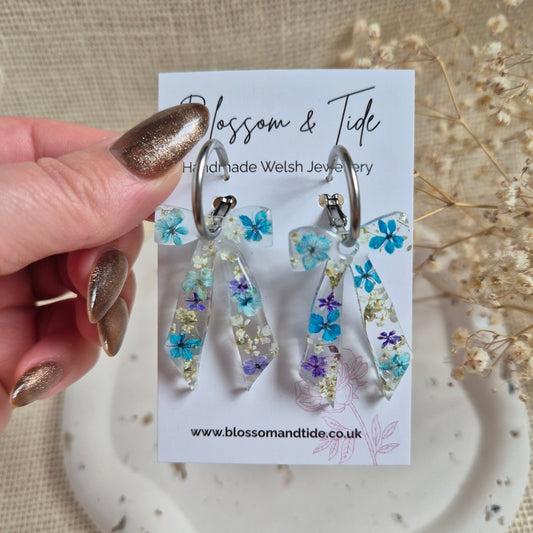 Bow earrings filled with a mix of blue and white pressed flowers on a stainless steel silver hoop. Presented on a Blossom & Tide branded card.