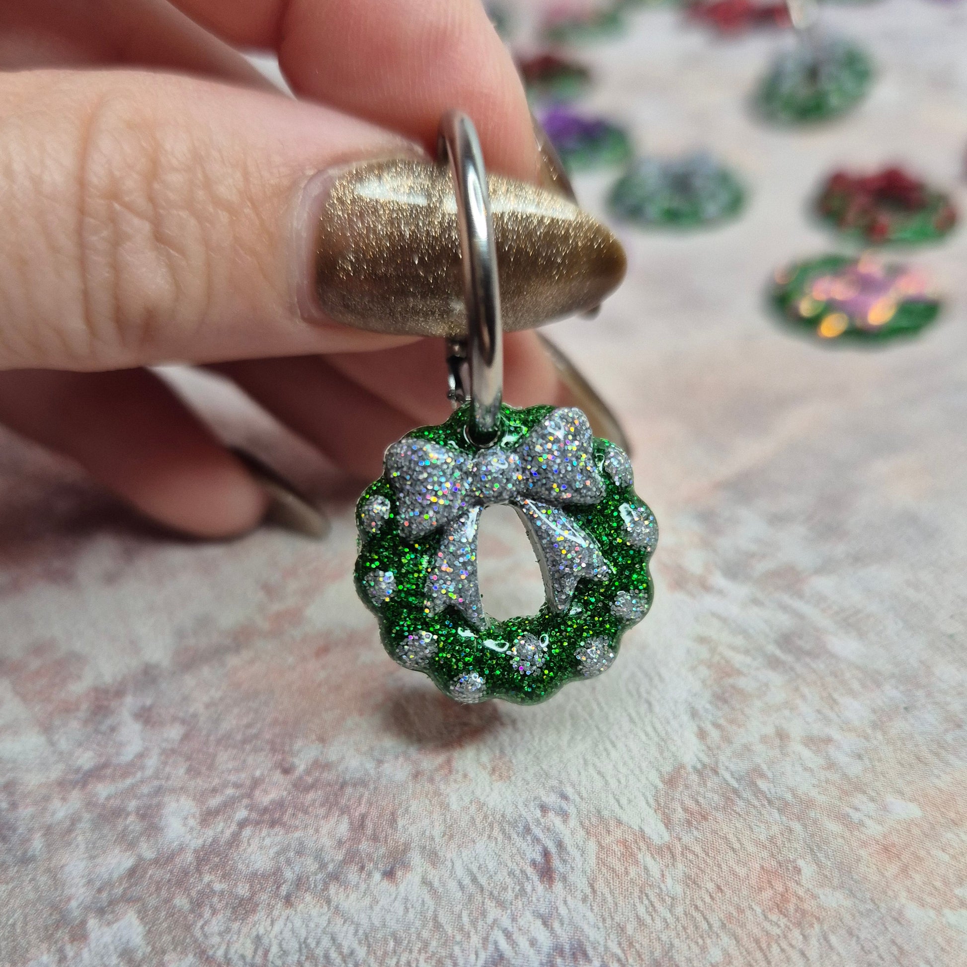 Christmas earrings with green and silver design held by a hand on a textured surface.
