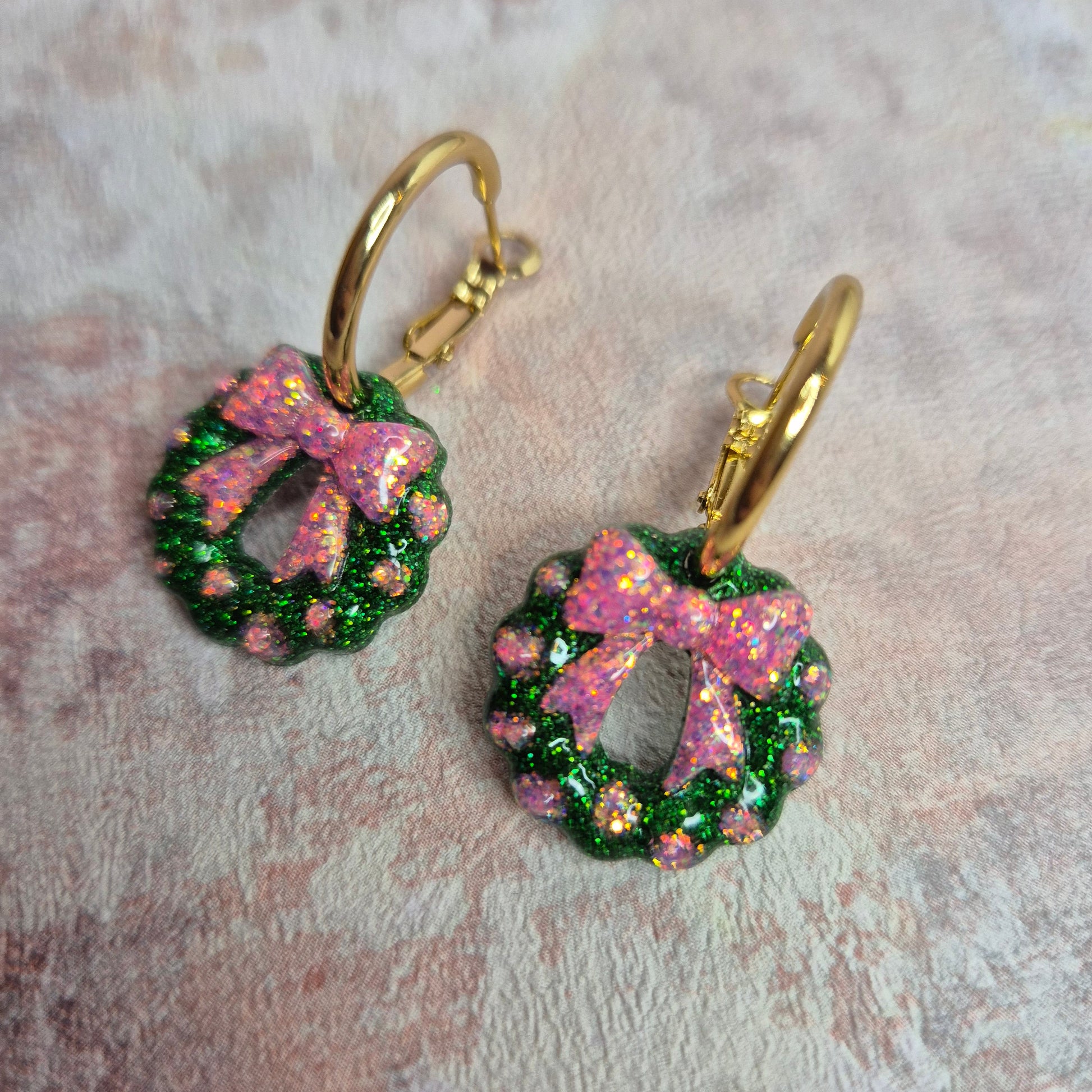 A Close up of a pair of Christmas hoop earrings in green and pink glitter. Wreath and bow design. Against a pink background