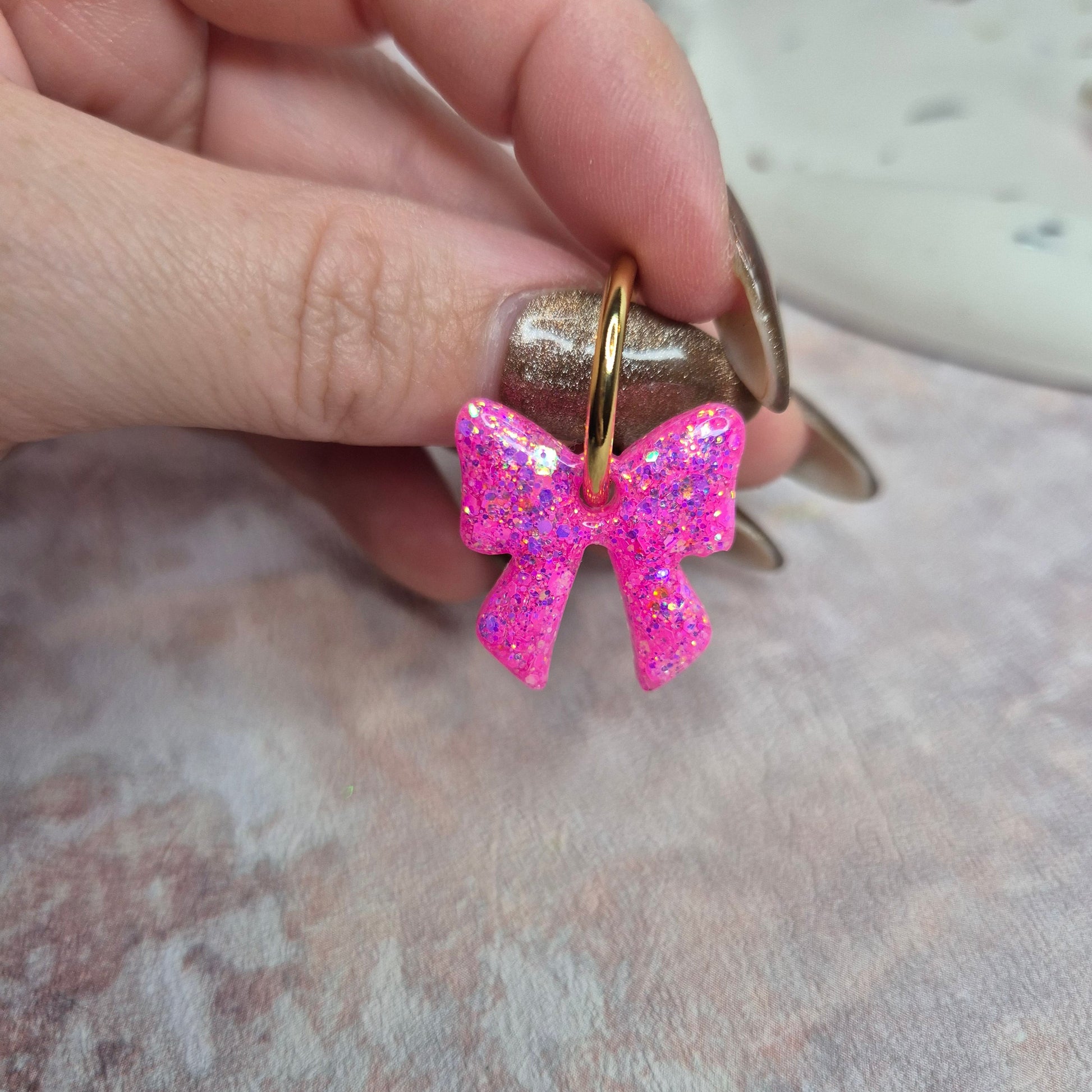 Hand holding a gold earring with a pink glittery bow on a textured surface