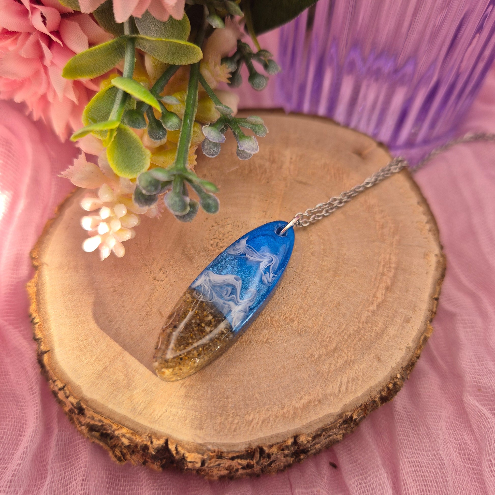 Handmade oval pendant necklace with real sand from Tenby beach, blue sea detail, and white wave ripples on a stainless steel chain.