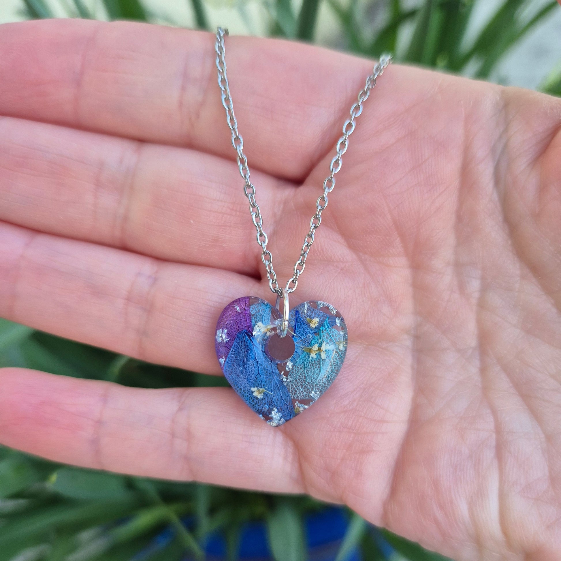 A heart-shaped resin necklace filled with blue and purple hydrangea petals, white flowers, and silver leaf on a silver-coloured stainless steel chain.