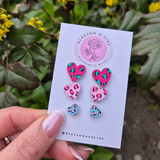 A set of three hand-painted heart-shaped stud earrings in bright leopard print designs, displayed on a branded card with an organza gift bag.