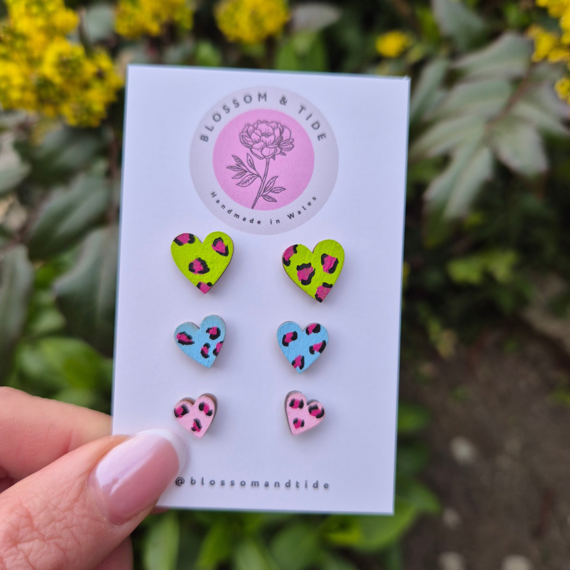 A set of three hand-painted heart-shaped stud earrings in bright leopard print designs, displayed on a branded card with an organza gift bag.
