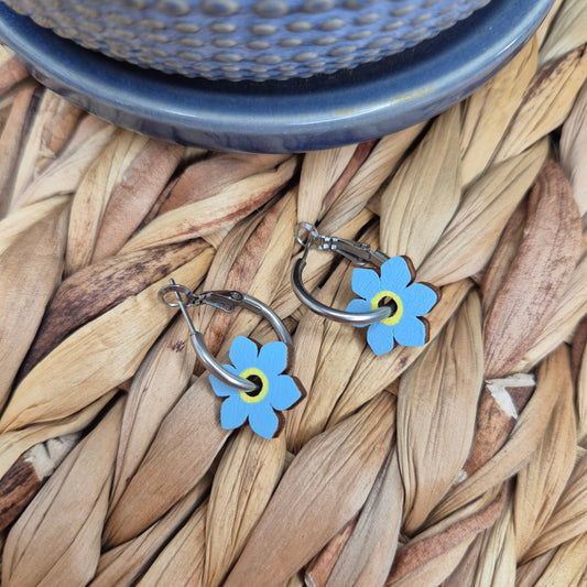 A pair of hand-painted hoop earrings featuring intricate forget-me-not flowers, sealed with a glossy resin topcoat, displayed on stainless steel hoops. Also available in a matte finish.