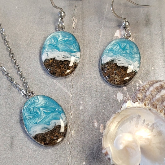 A jewelry set featuring a silver chain necklace and matching drop earrings with oval resin pendants. The pendants depict a wave design with light blue and white swirling on the top half and dark brown sand on the bottom half, styled on a gray textured surface with a white clam shell.