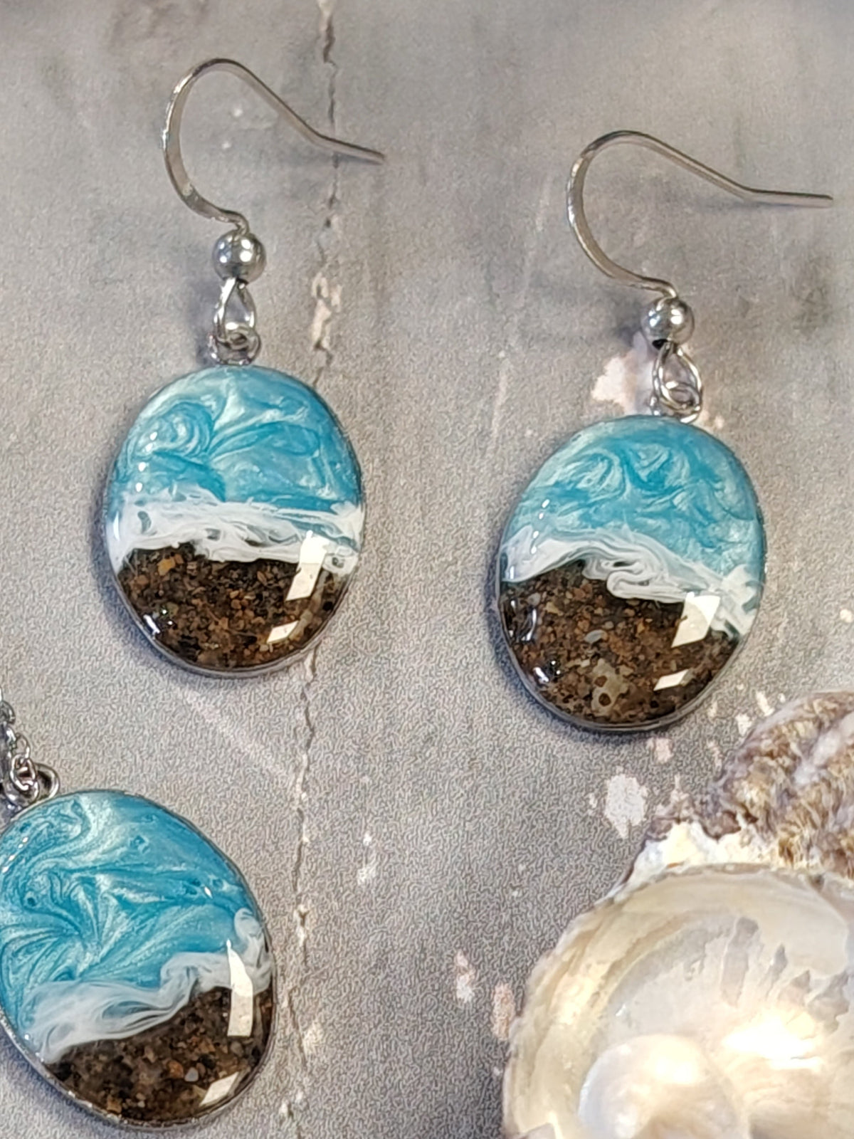 A jewelry set featuring a silver necklace and matching drop earrings with oval charms. The charms depict a stylized beach scene with light blue and white waves on top and brown sand on the bottom, styled on a gray textured surface with seashells.