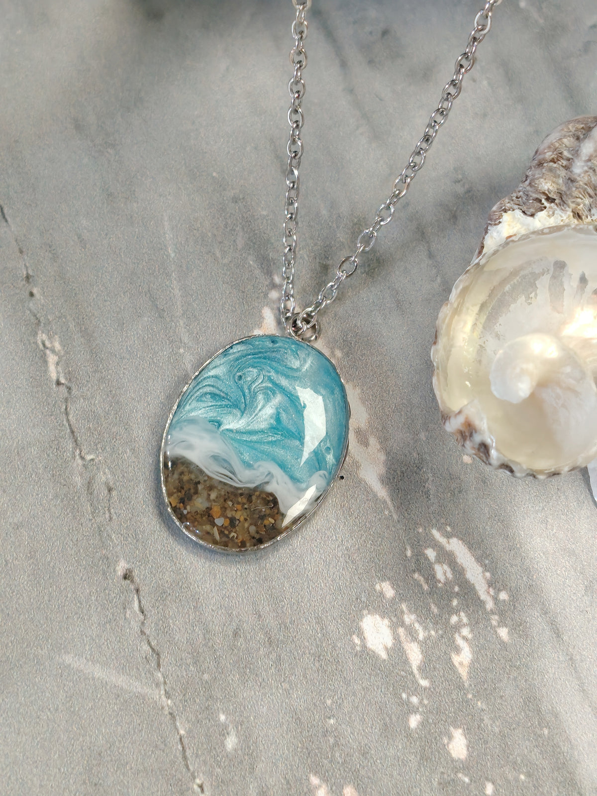 An oval pendant necklace with a silver chain, featuring a resin design of a blue ocean wave and beach sand, displayed on a gray stone surface next to a seashell.