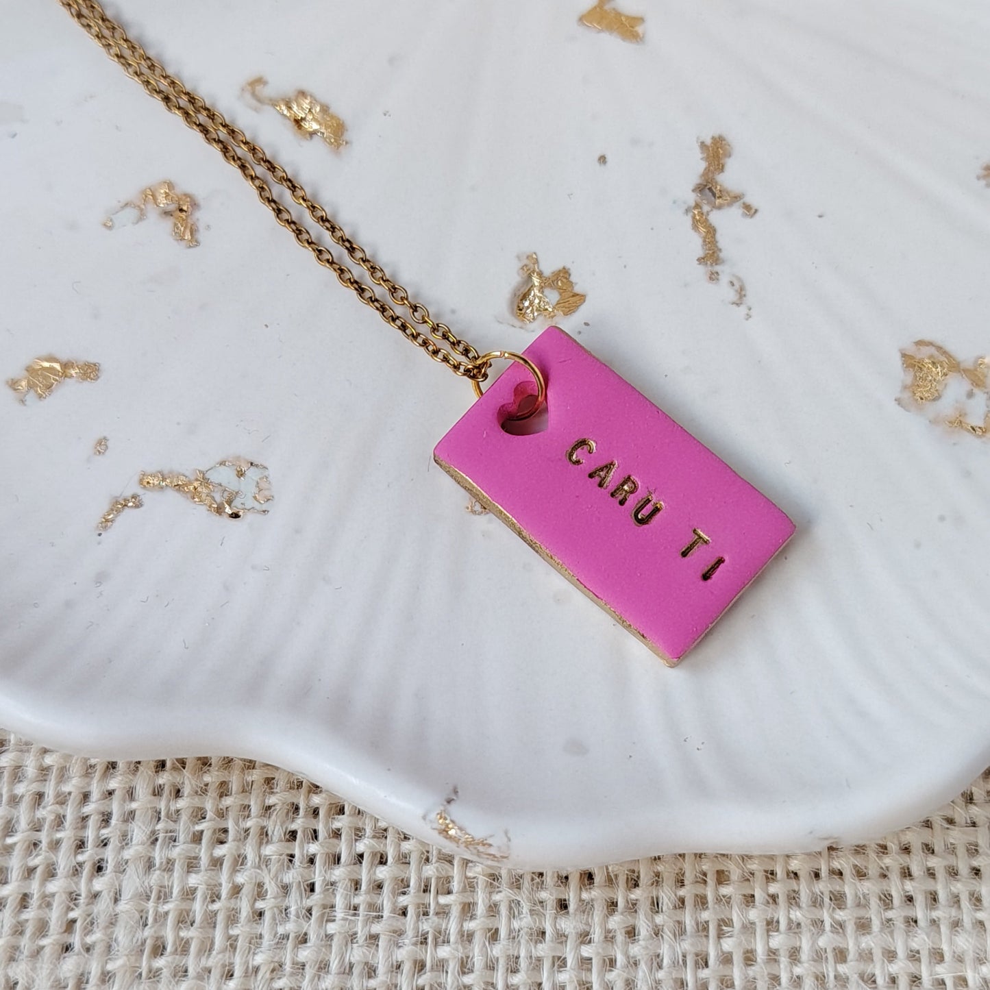 Pink polymer clay rectangle necklace with gold hand-painted accents, heart cut-out and the Welsh words Caru Ti on a gold stainless steel chain. Against a neutral background with gold flecks.