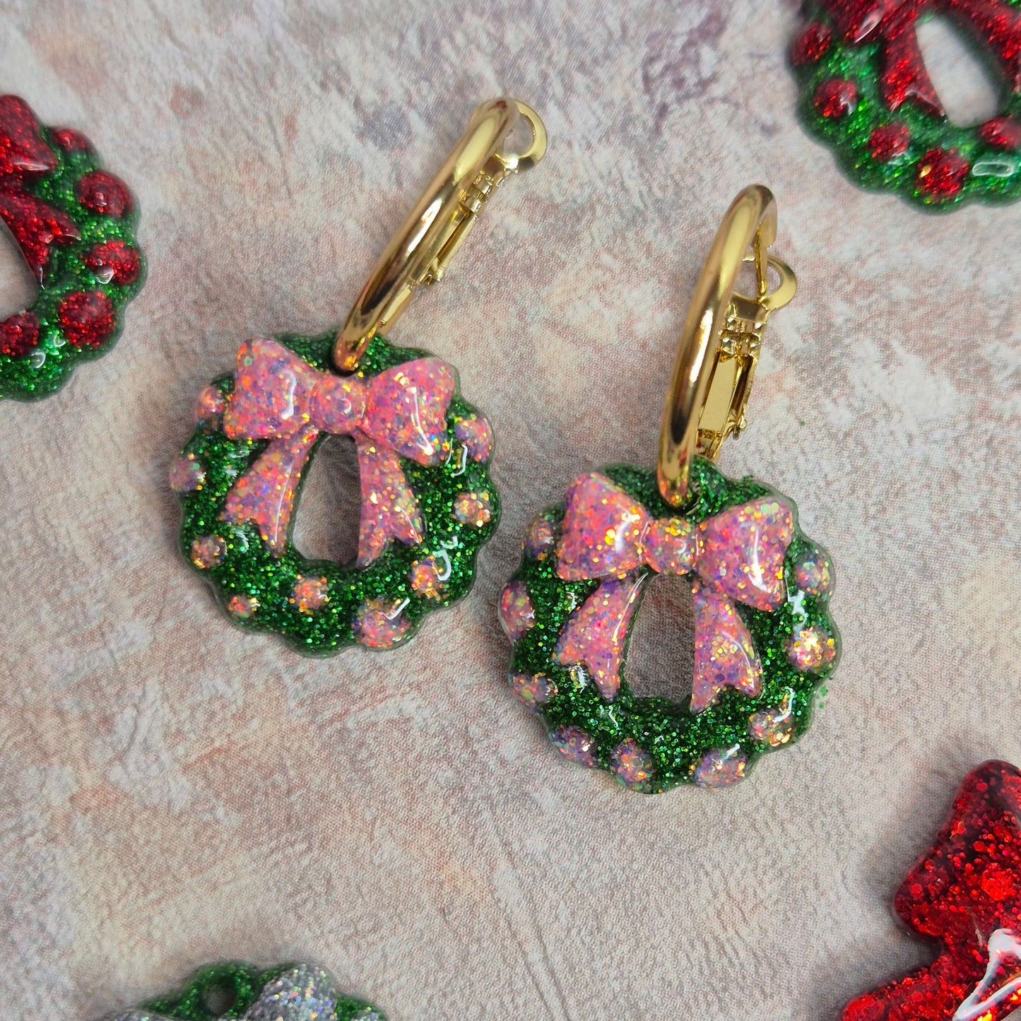 Pink and green glitter wreath earrings on gold hoop against a pink background. 