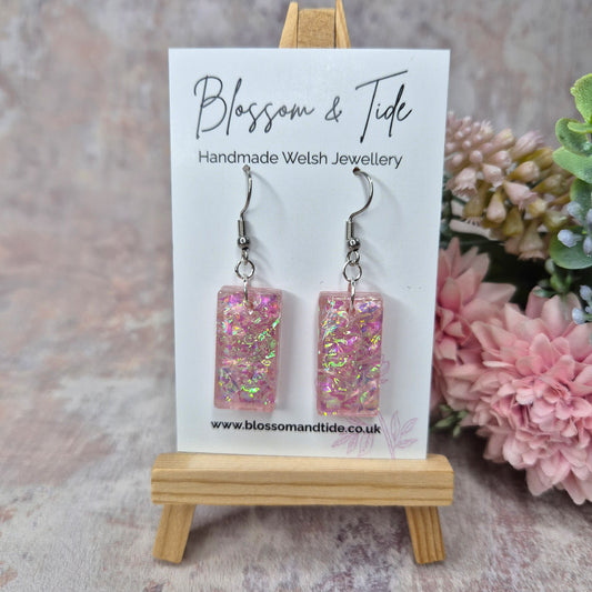 Pink dichroic resin rectangular earrings on a 'Blossom & Tide' card with flowers in the background.