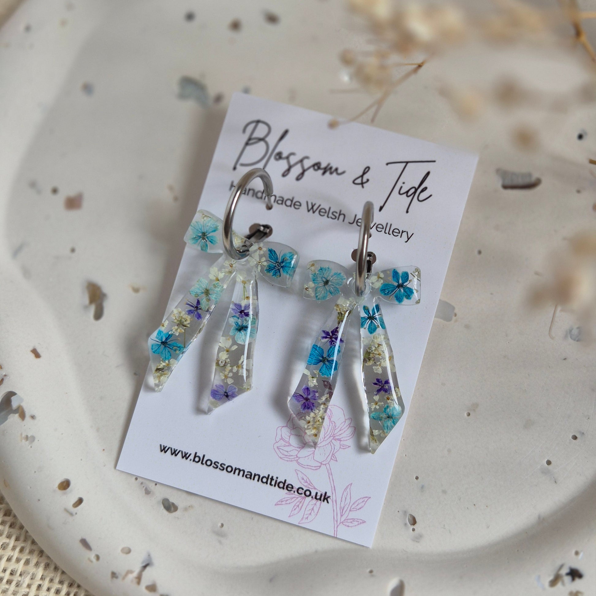 Bow earrings filled with a mix of blue and white pressed flowers on a stainless steel silver hoop. Presented on a Blossom & Tide branded card.