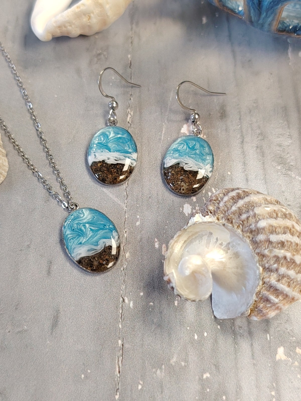 A jewelry set featuring a silver chain necklace and matching drop earrings with oval resin pendants. The pendants depict a wave design with light blue and white water, dark brown sand, and a silver fishhook ear wire.