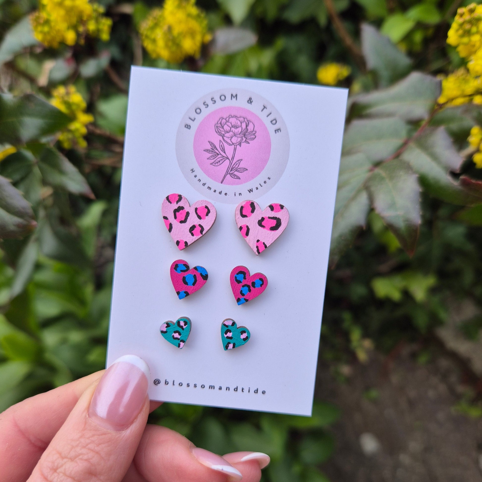 A set of three hand-painted heart-shaped stud earrings in bright leopard print designs, displayed on a branded card with an organza gift bag.