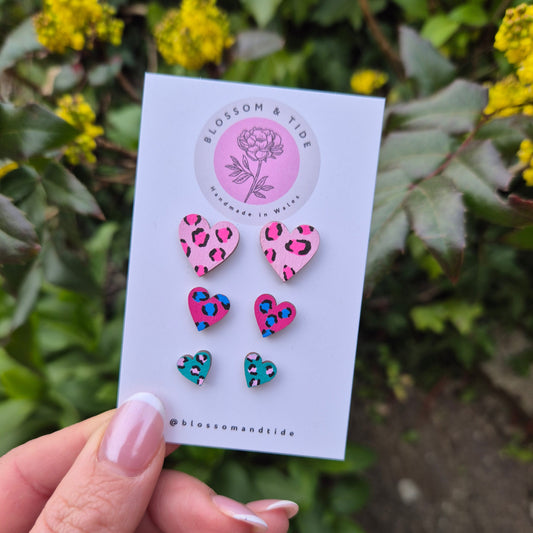 A set of three hand-painted heart-shaped stud earrings in bright leopard print designs, displayed on a branded card with an organza gift bag.