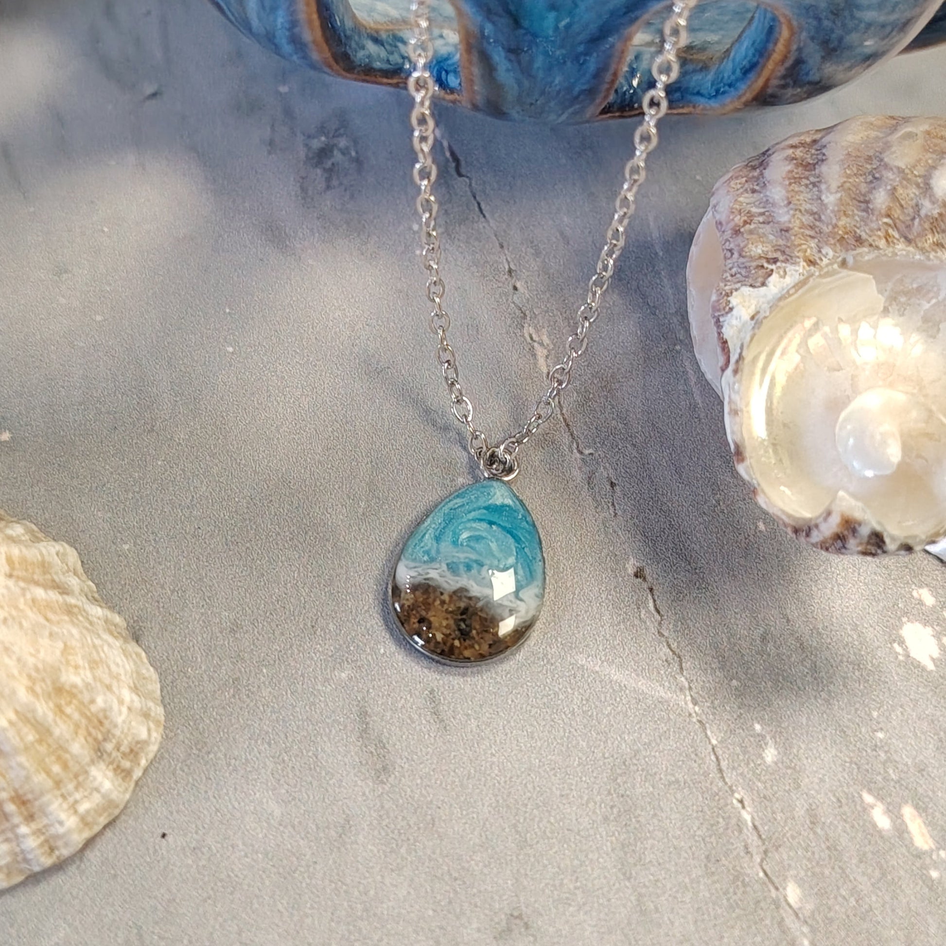 A silver chain necklace with a teardrop-shaped pendant containing a swirl of turquoise blue and white resin with sand inclusions, displayed on a gray surface with seashells and a blue ceramic object in the background.