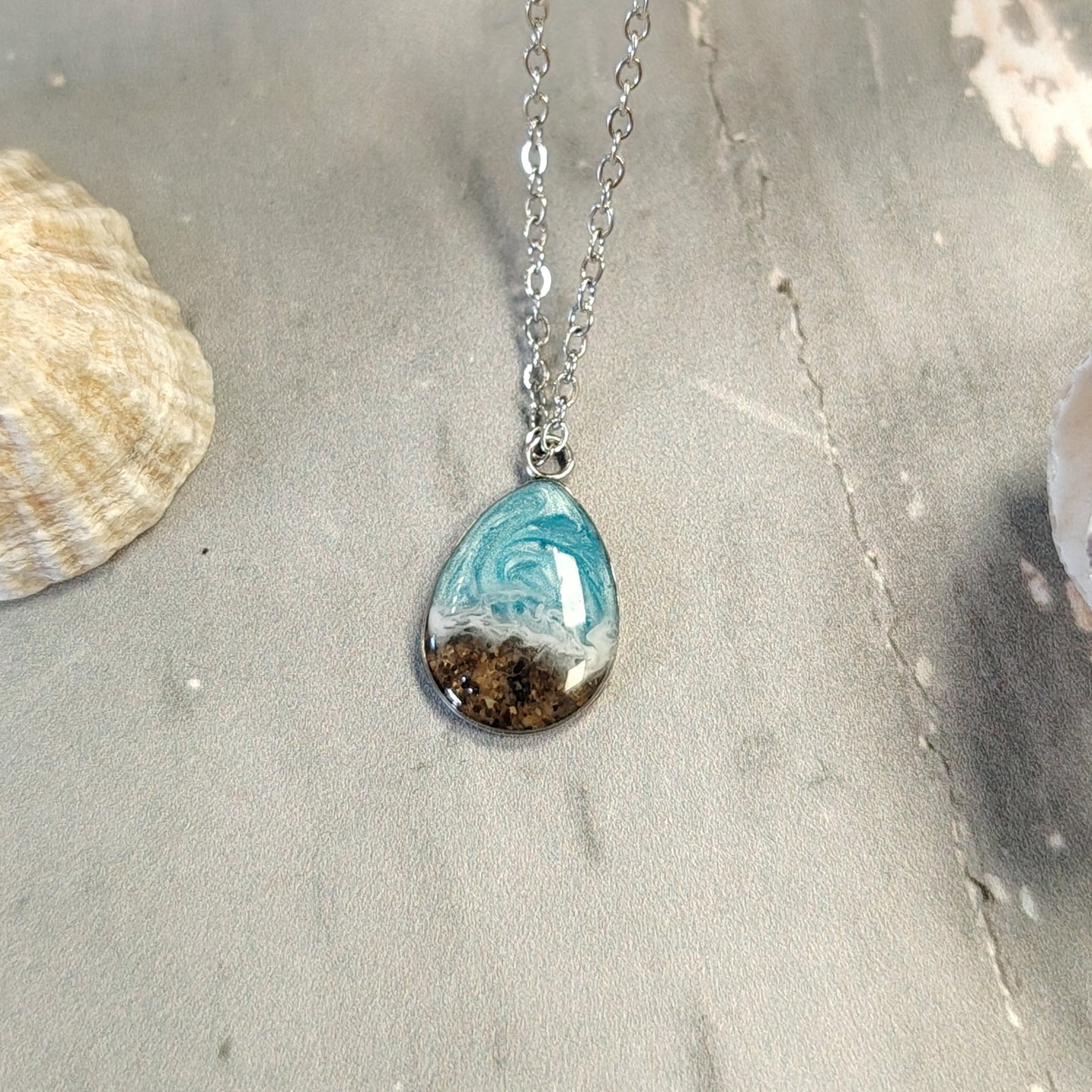 A silver chain necklace with a teardrop-shaped pendant containing a swirl of turquoise blue and white resin with sand inclusions, displayed on a gray textured surface with a seashell prop.