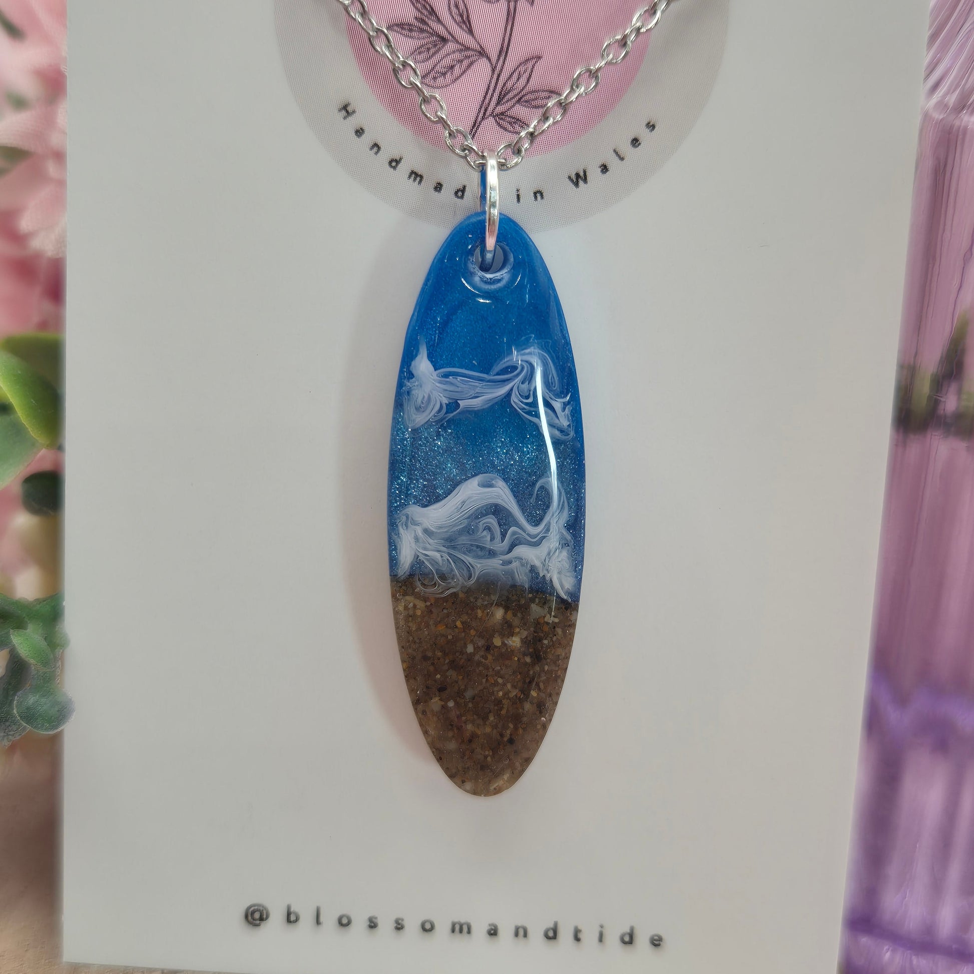 Close-up of a long oval beach-inspired necklace featuring golden sand, blue resin, and white wave-like ripples, made in Wales.