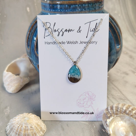 A teardrop-shaped blue sea pendant  with real Welsh sand necklace on a silver chain, displayed on a white card labeled 'Blossom & Tide Handmade Welsh Jewellery'. The card is styled on a gray wooden surface with seashells and a blue ceramic vase in the background.