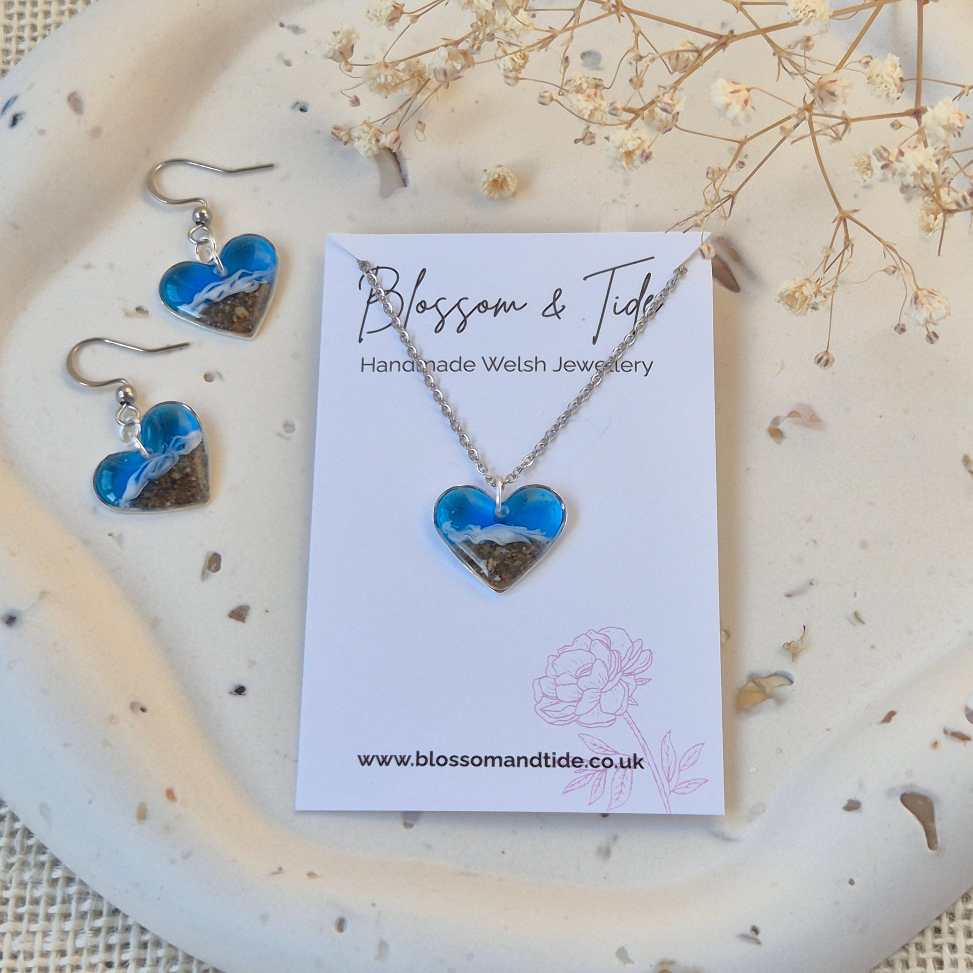 heart shaped beach necklace and earrings on a branded card, placed on a textured surface with dried flowers.