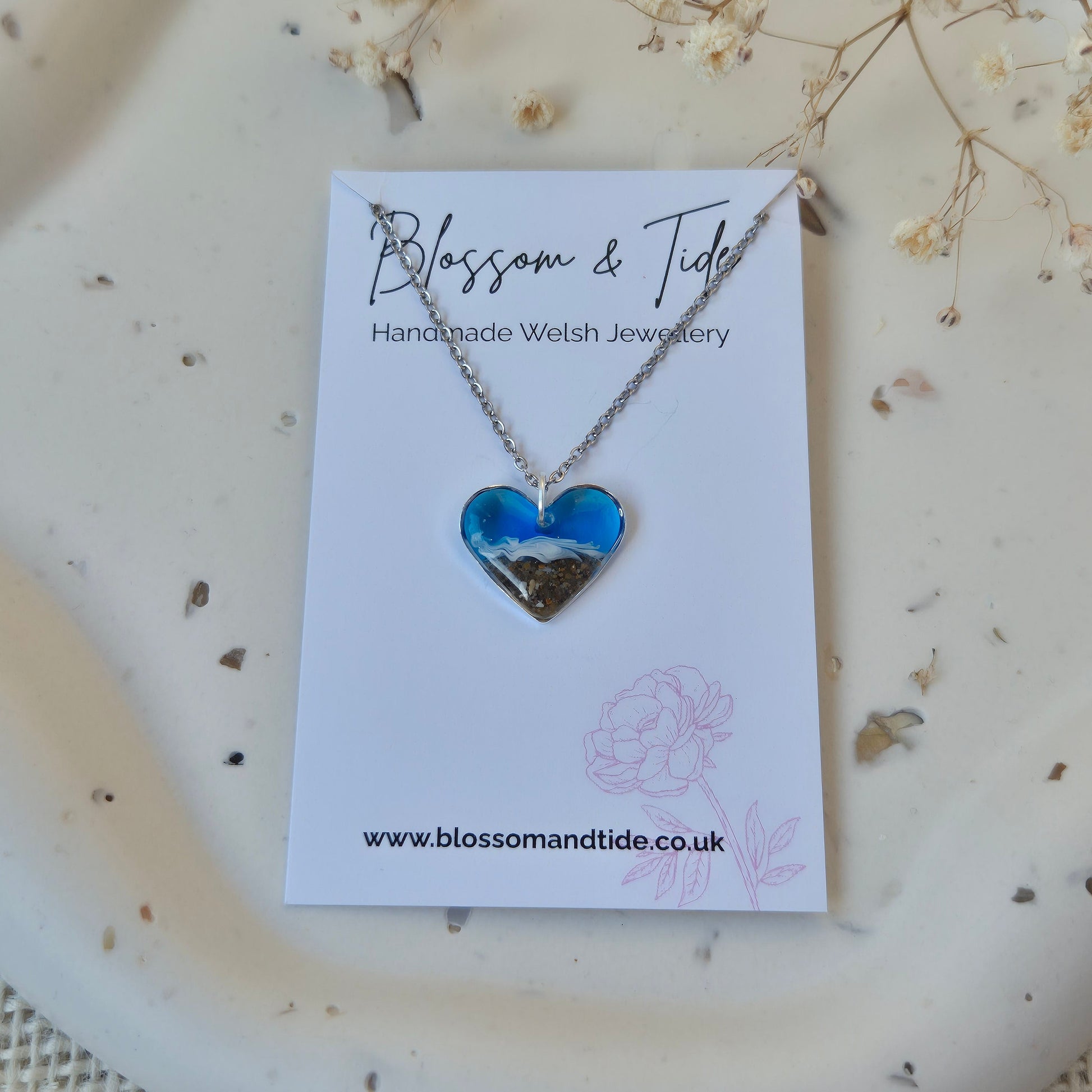 Necklace with blue heart beach pendant on a branded card, placed on a textured surface with dried flowers.