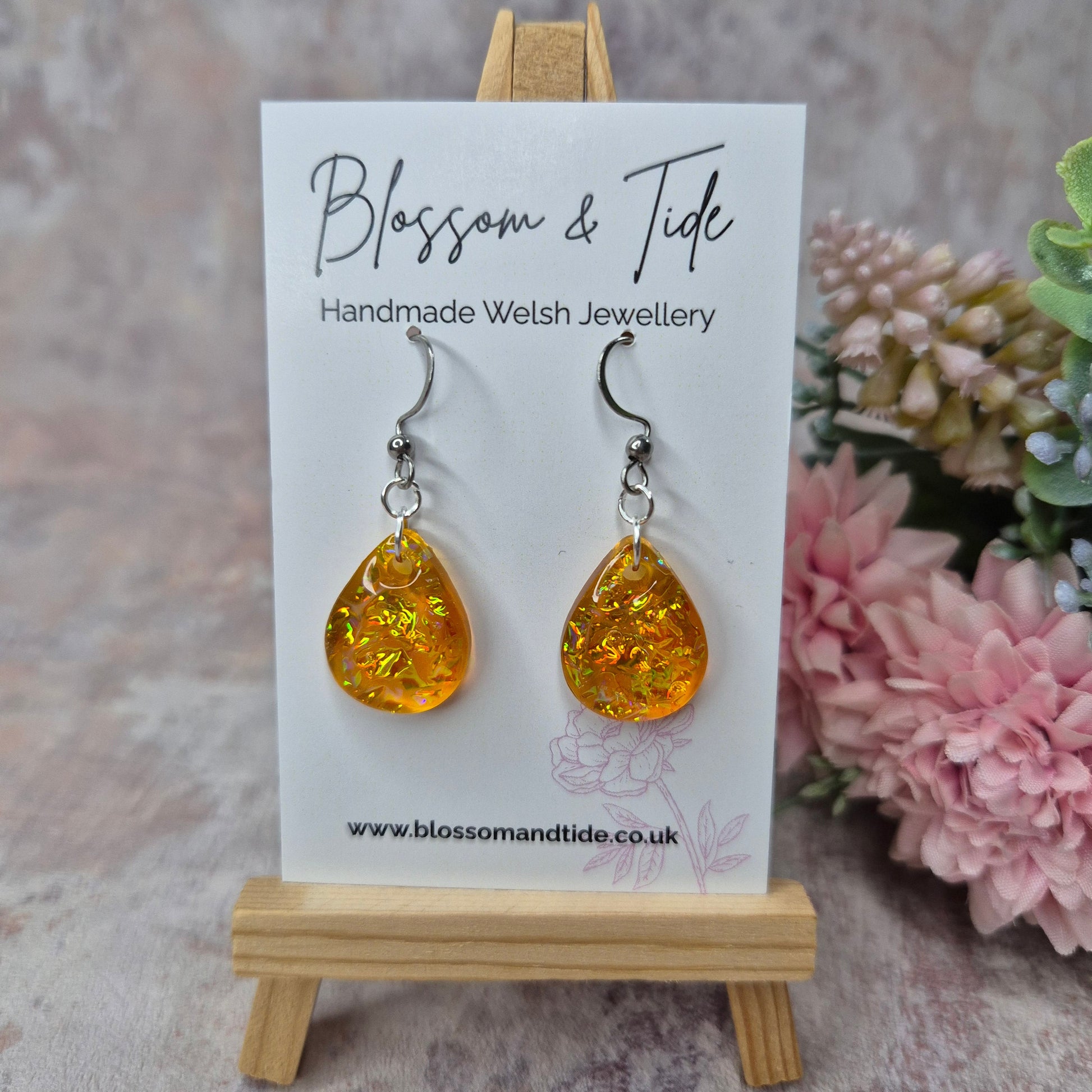 Teardrop shape orange dichroic resin earrings on branded card, against a pink background