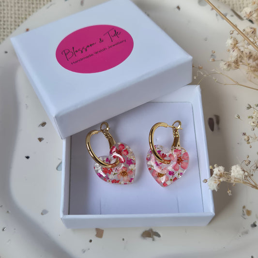 Floral earrings in a white box with a pink blossom and tide label on a textured surface.