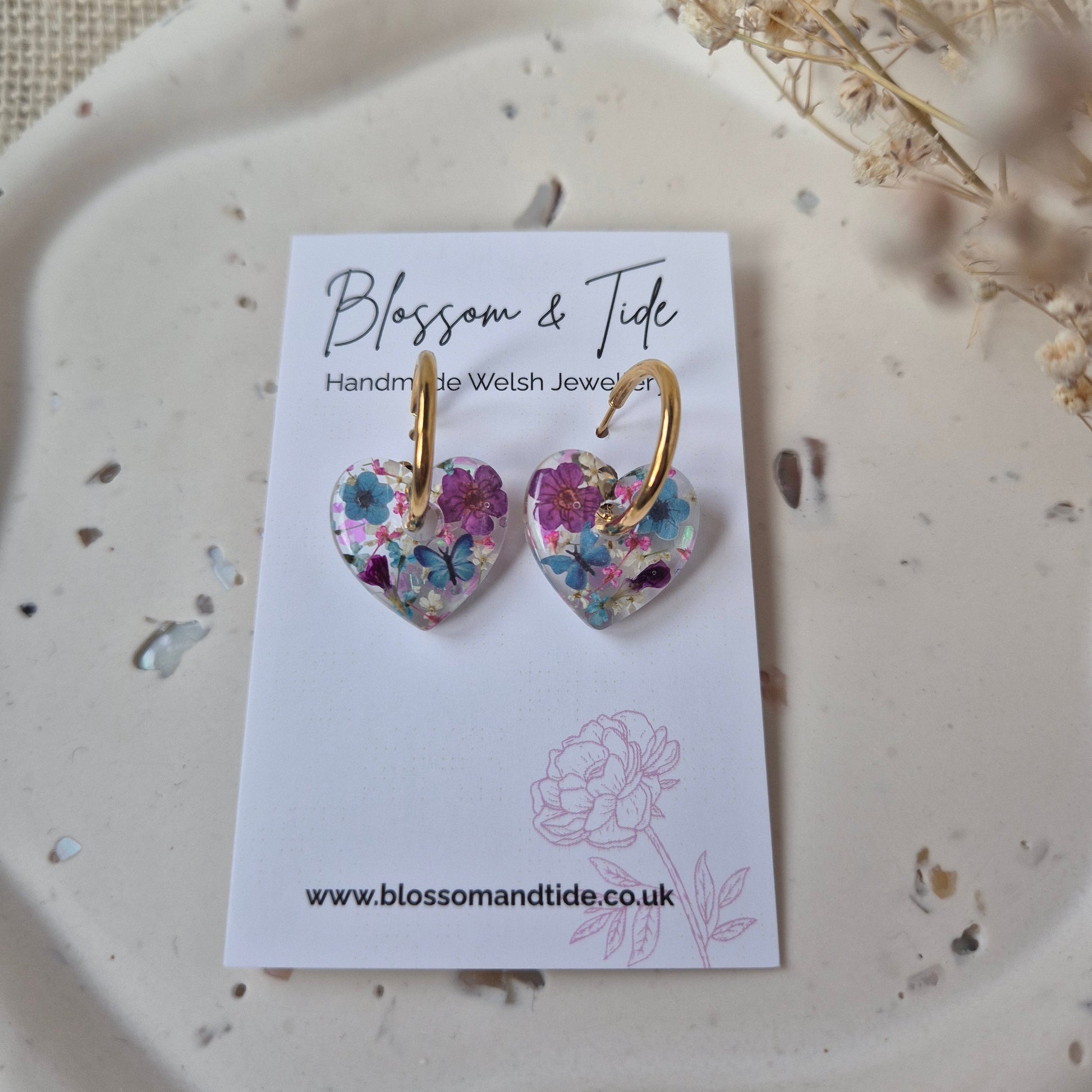 Floral earrings on a card with 'Blossom & Tide' branding, placed on a textured surface.