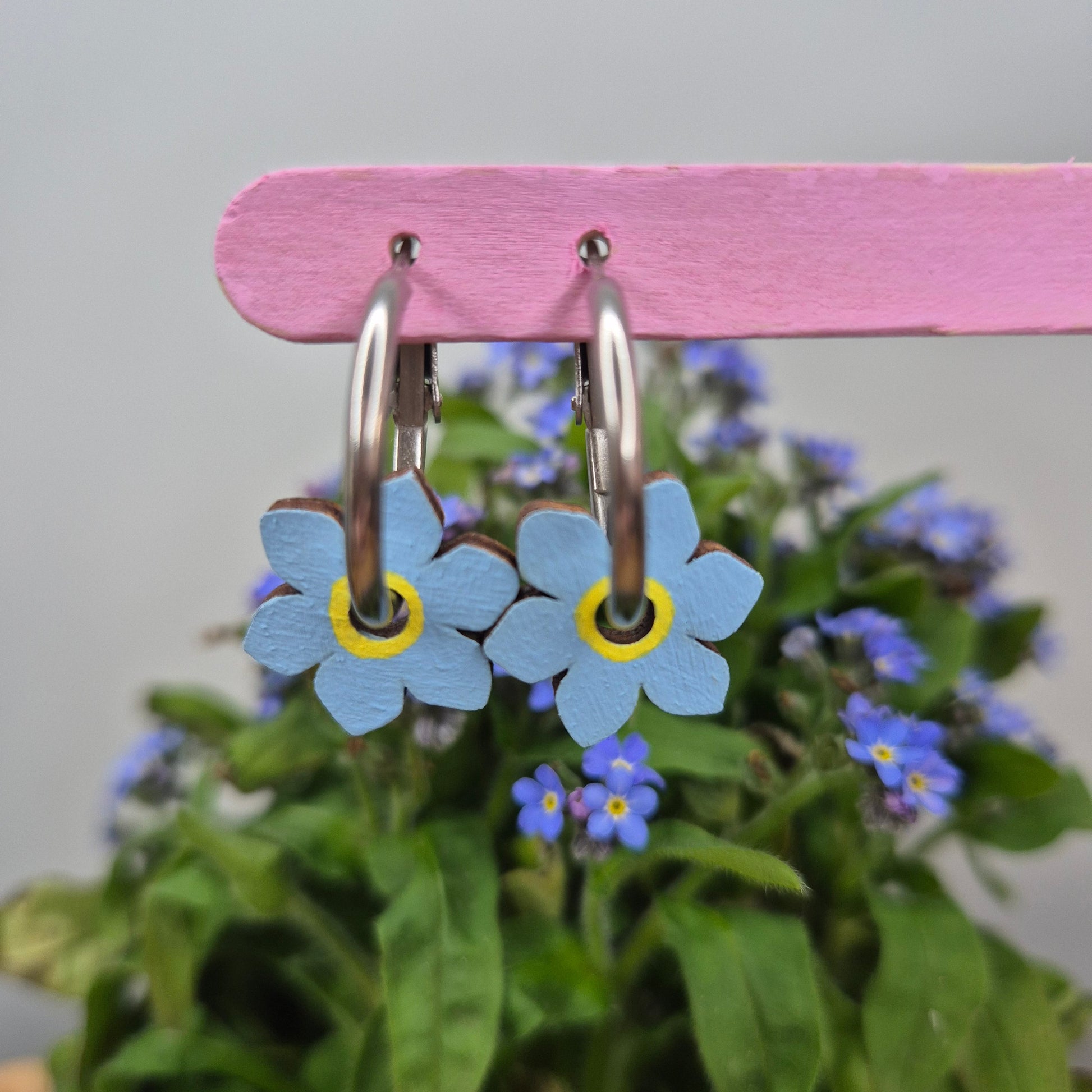 A pair of hand-painted hoop earrings featuring intricate forget-me-not flowers, sealed with a glossy resin topcoat, displayed on stainless steel hoops. Also available in a matte finish.