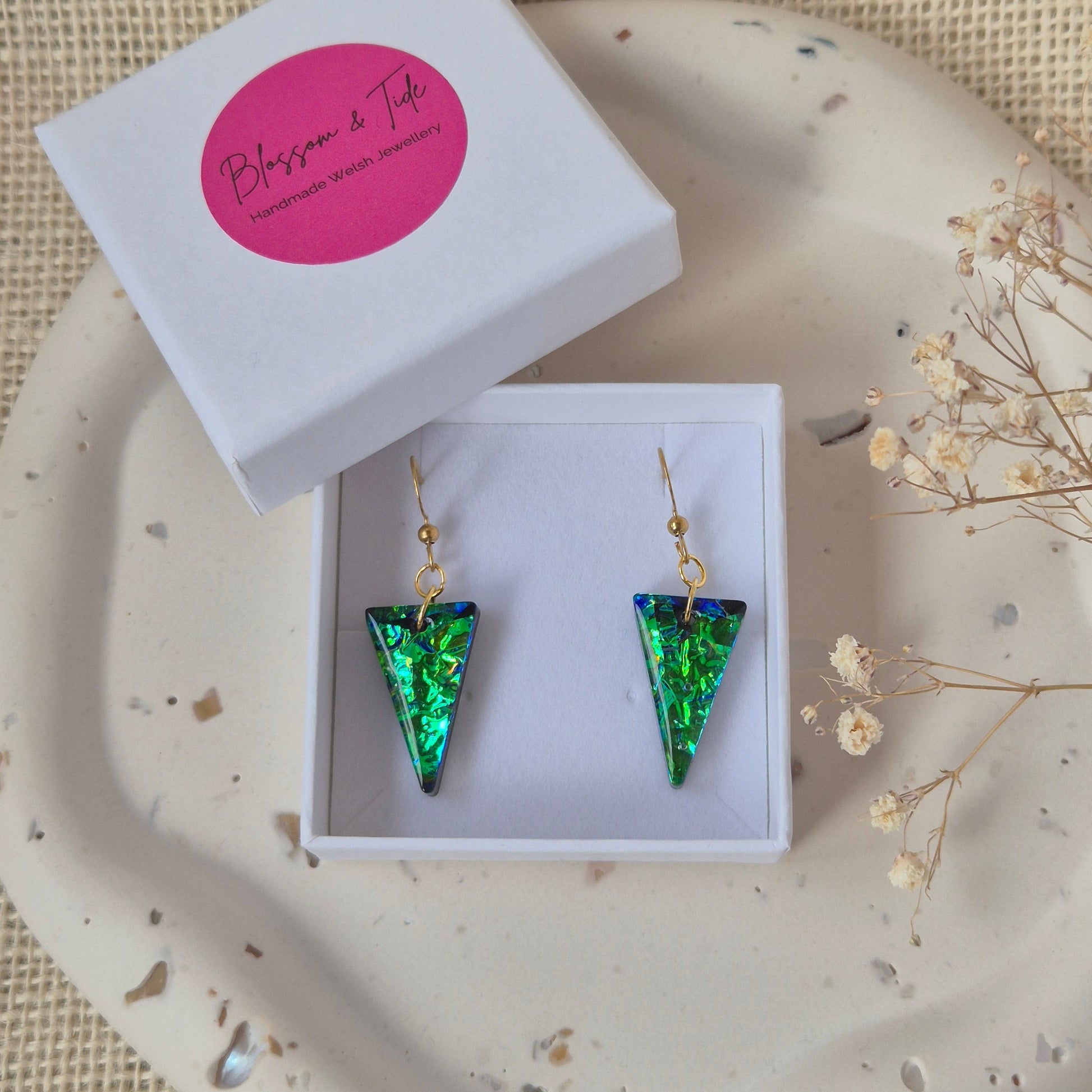 Green and black triangle-shaped earrings in a white box on a textured surface with dried flowers.