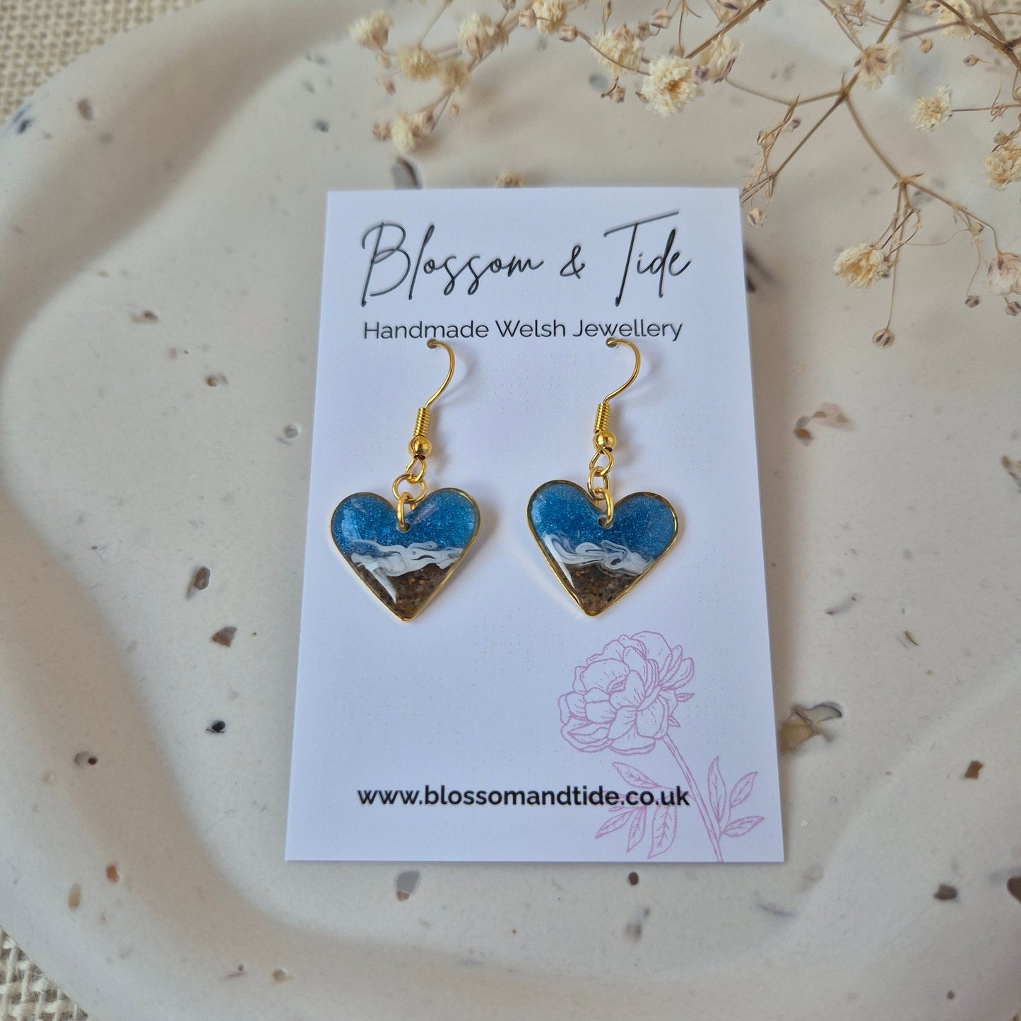 Heart-shaped sand earrings on a card with 'Blossom & Tide' branding on a textured surface.