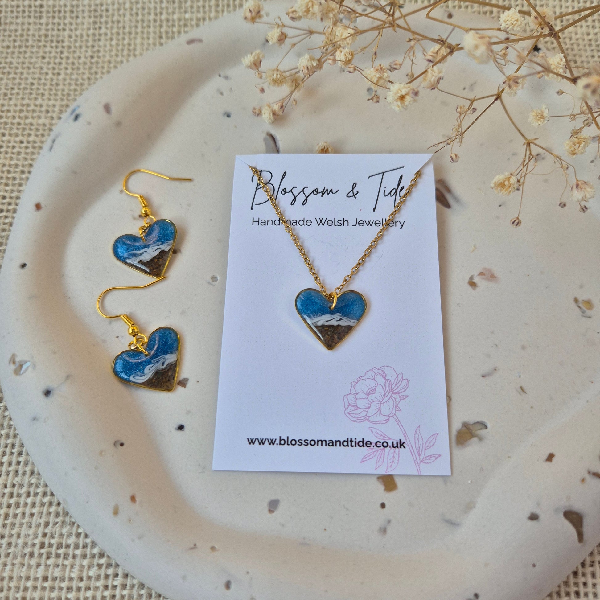 heart shaped sand earrings and matching necklace with gold accents on a branded card, displayed on a textured surface.