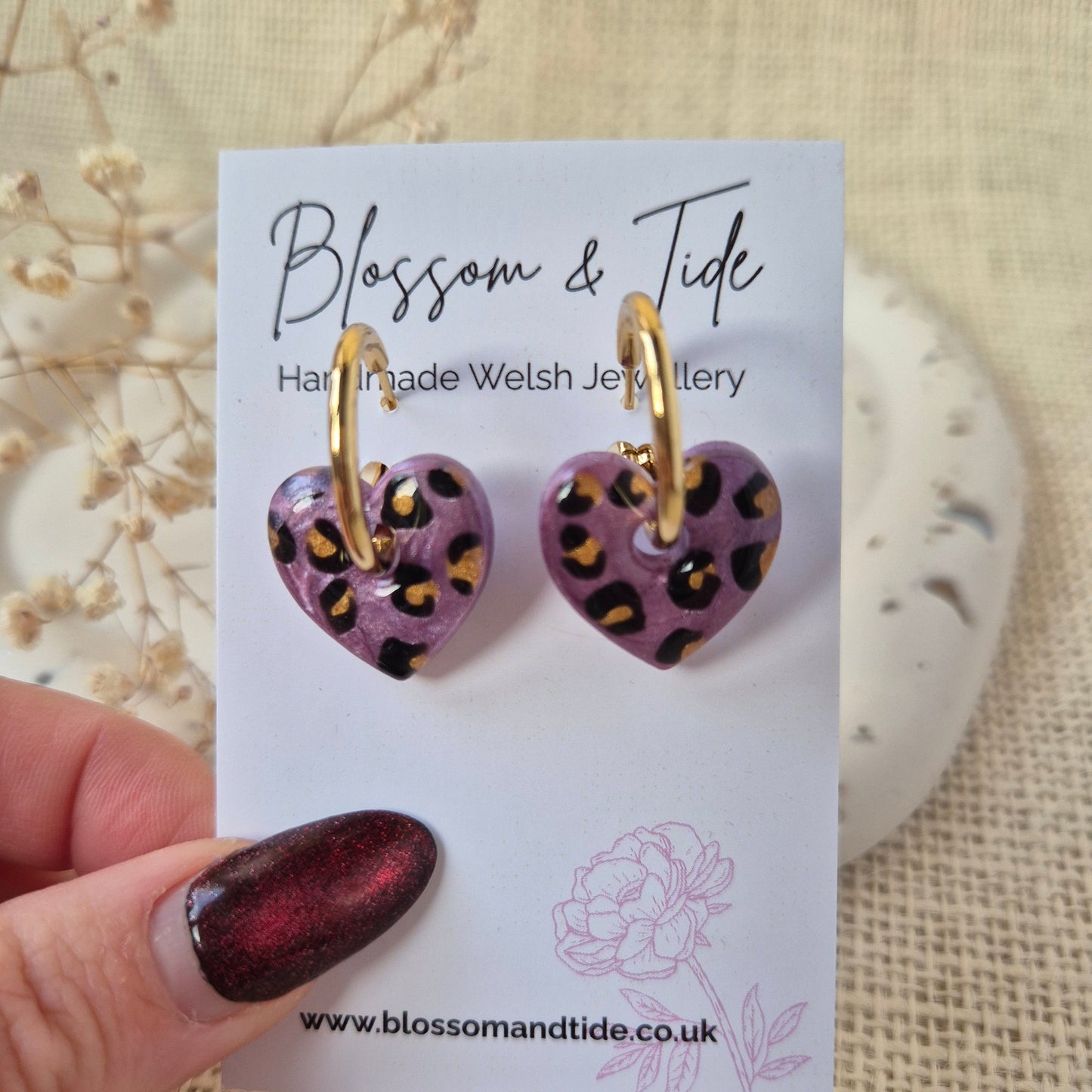 Pair of leopard heart earrings on a 'Blossom & Tide' card held by a hand with dark nail polish.