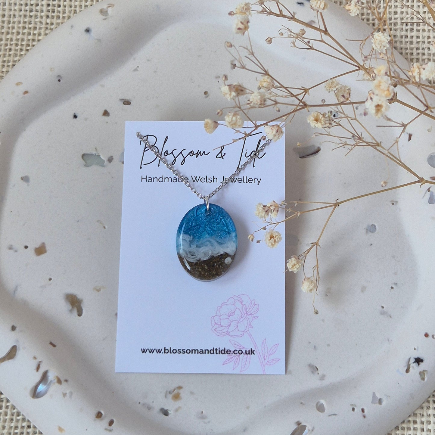 Necklace with a blue and white pendant on a card, placed on a textured surface with dried flowers.