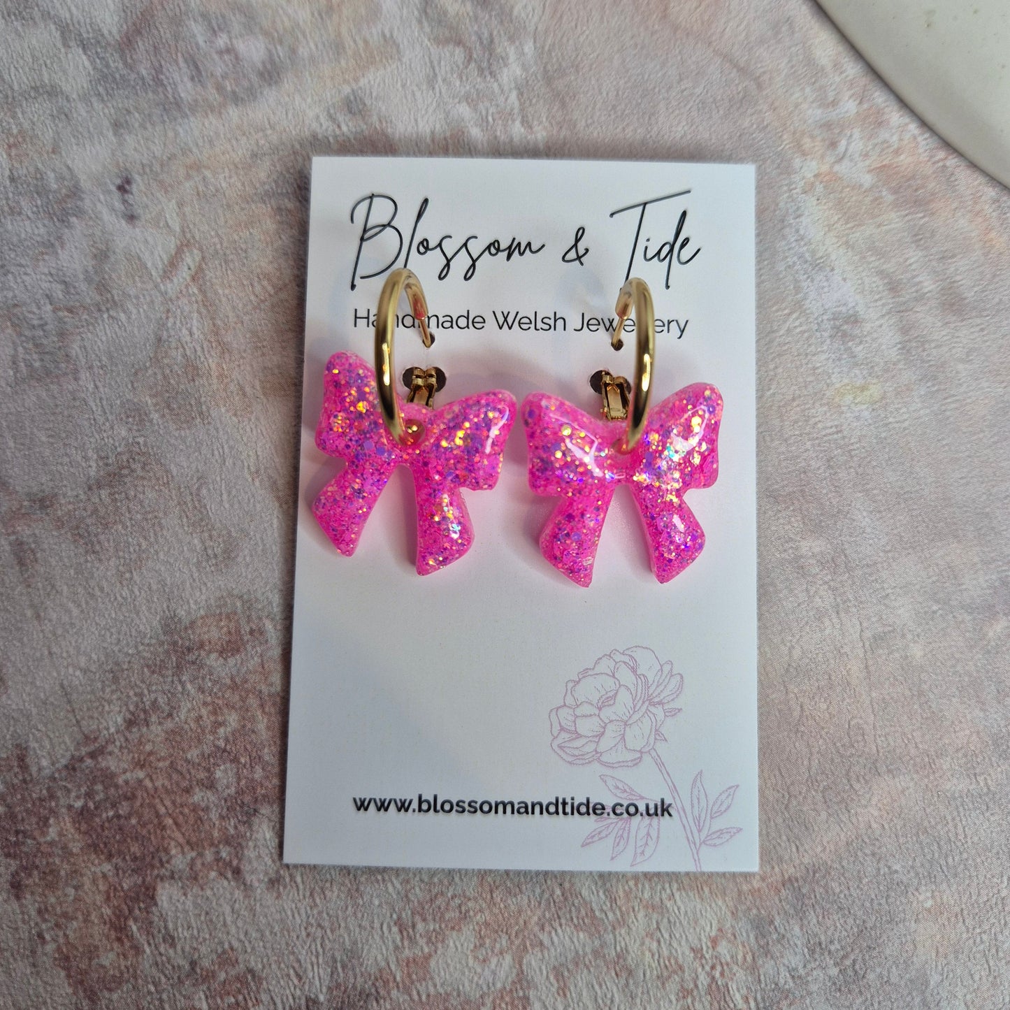 Pink bow-shaped earrings on a branded card with a marble background
