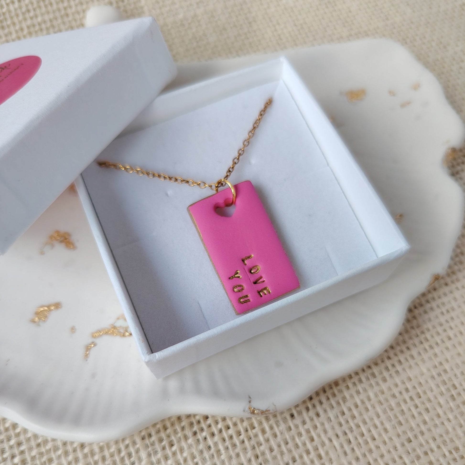 Pink clay necklace reading 'LOVE YOU' in a white box on a textured beige surface