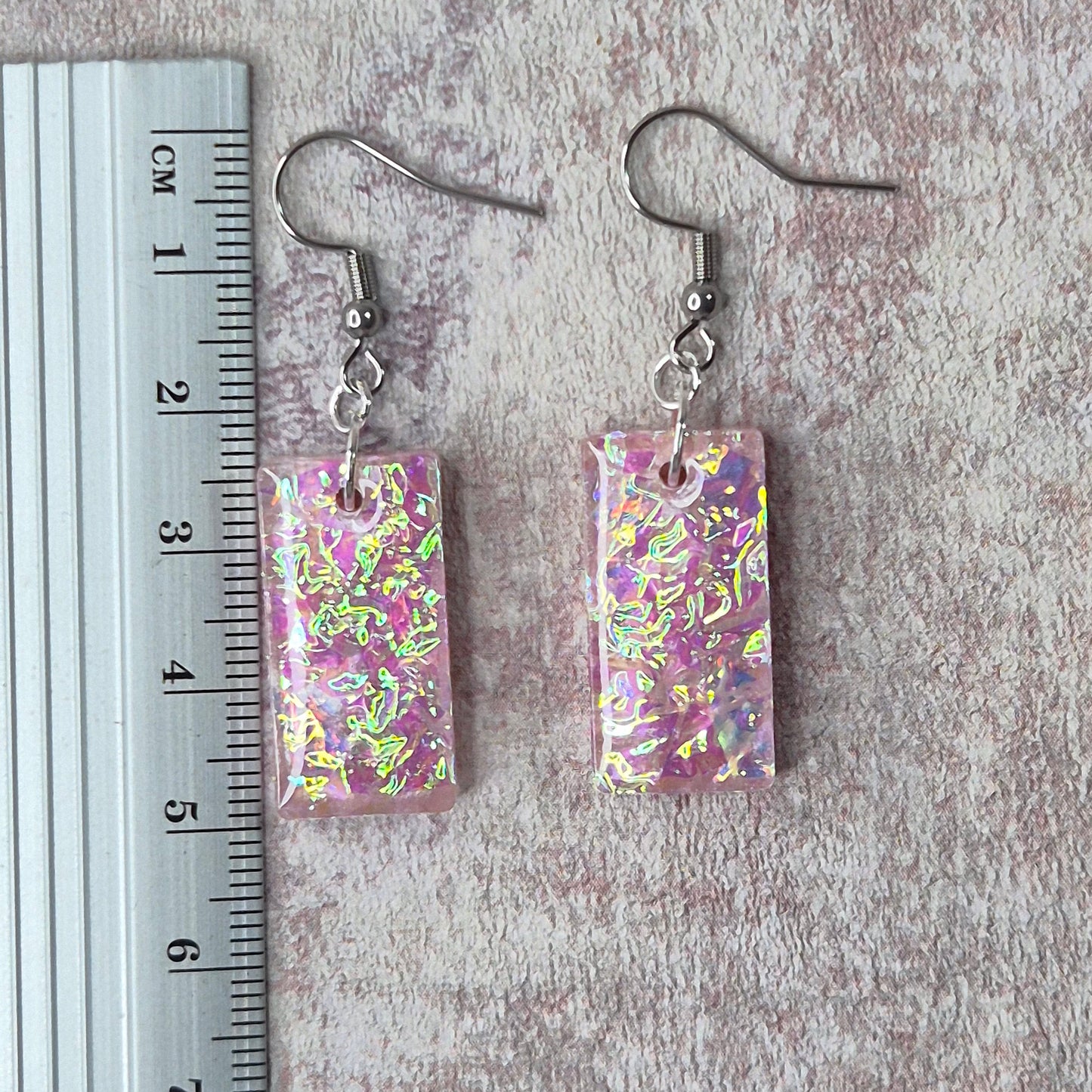 Pair of pink dichroic resin earrings  on a pink textured surface with a ruler for scale.