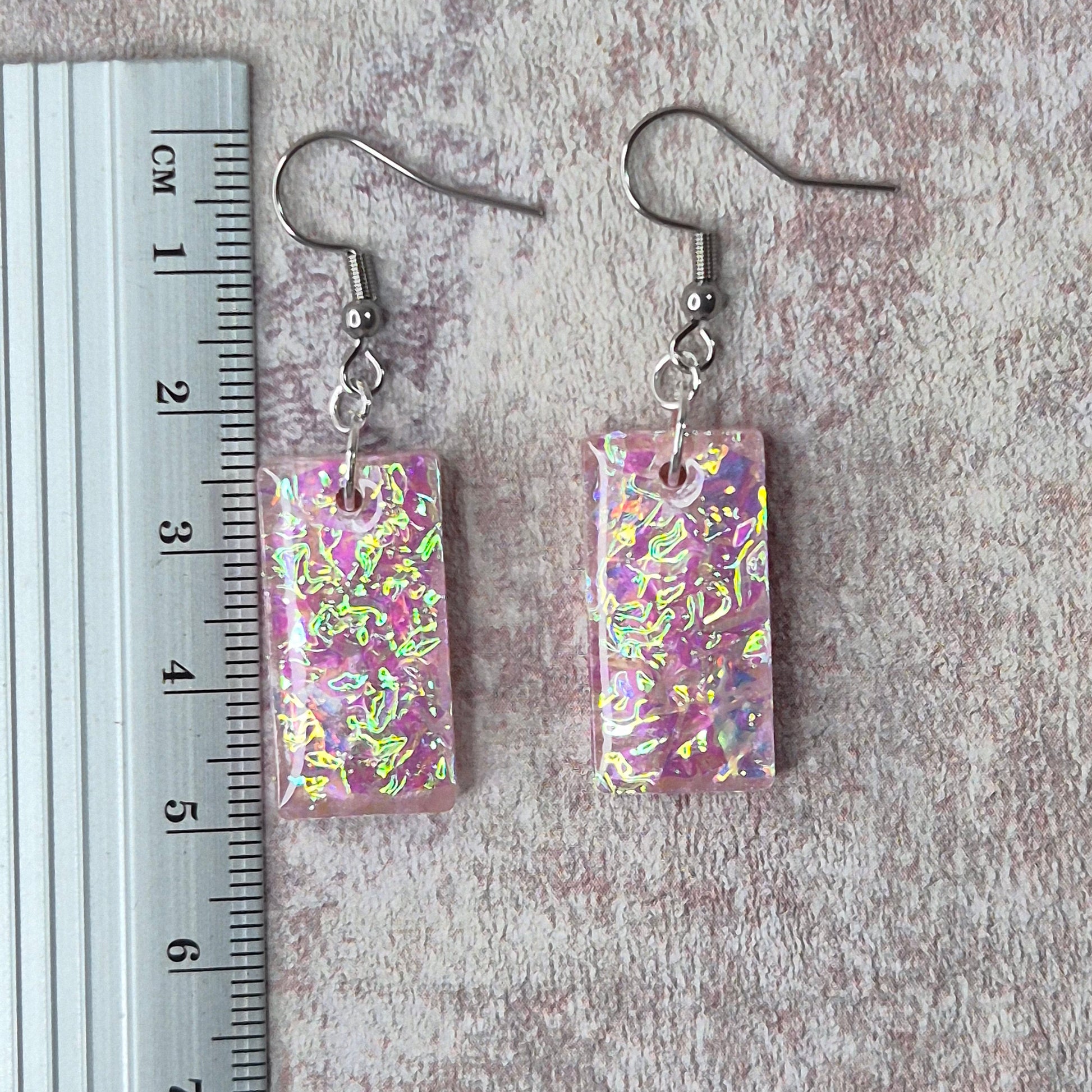 Pair of pink dichroic resin earrings  on a pink textured surface with a ruler for scale.