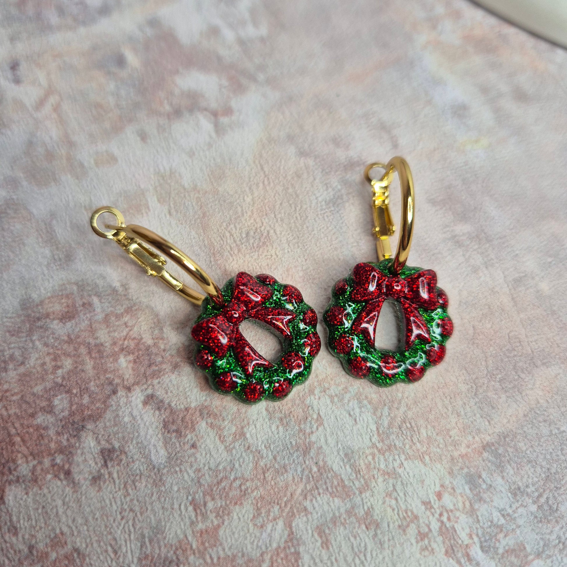 Red and green wreath-shaped earrings with gold hoops on a textured surface