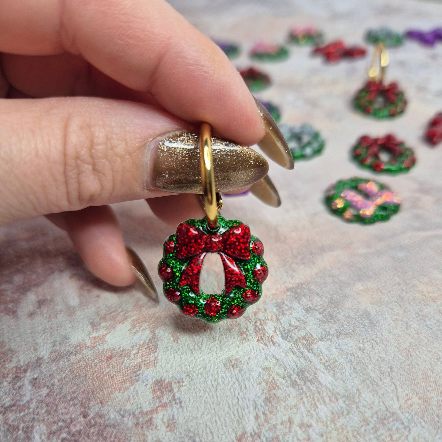Hand holding a gold hoop earring with a red and green wreath-shape against a textured background